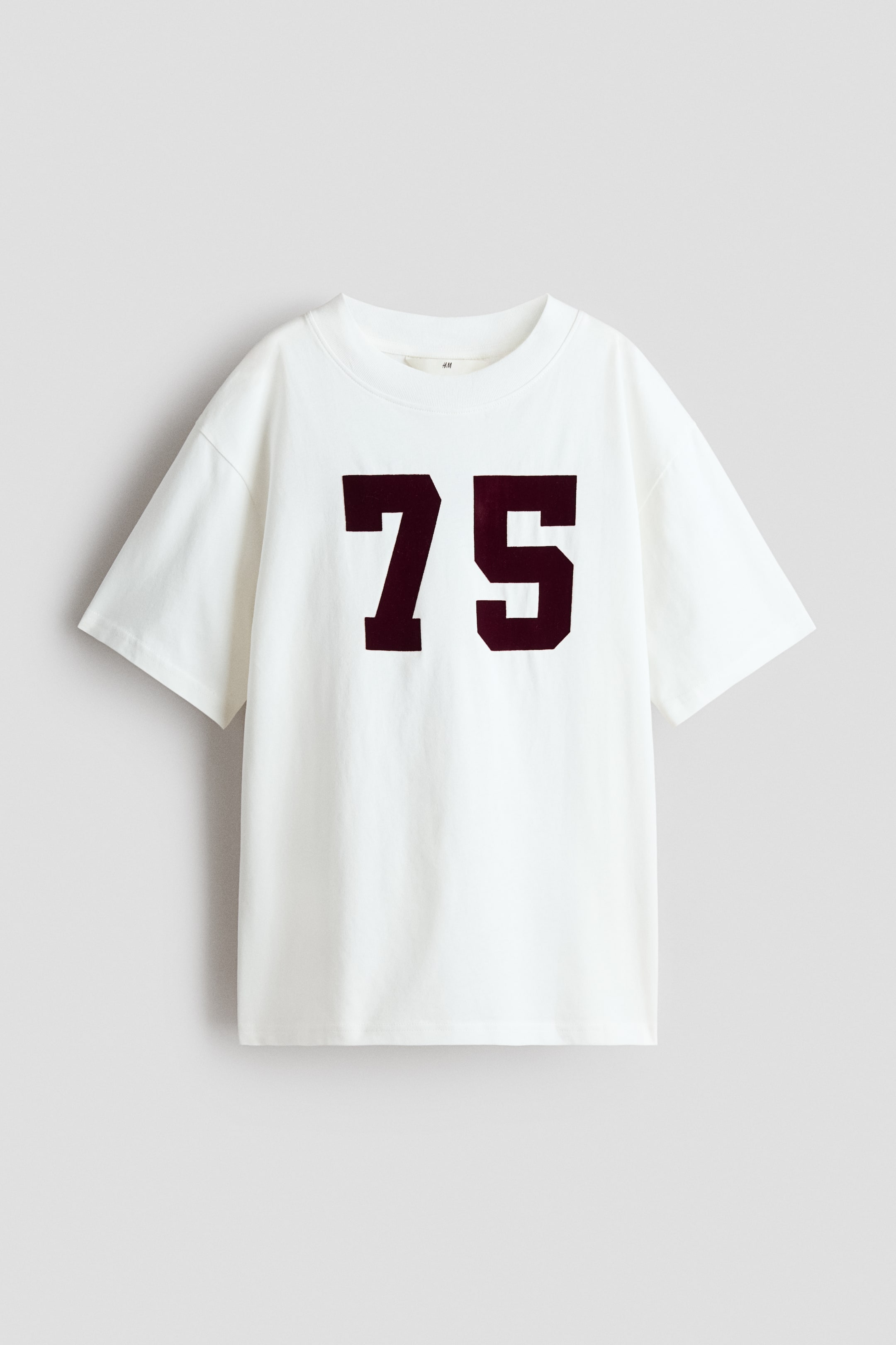 Προβολή μεγαλύτερης εικόνας: Ένα λευκό T-shirt φαίνεται από μπροστά. Φέρει έναν μεγάλο, σκούρο μπορντό «75» στο κέντρο του στήθους και έχει στρογγυλή ριμπ λαιμόκοψη και κοντά μανίκια.