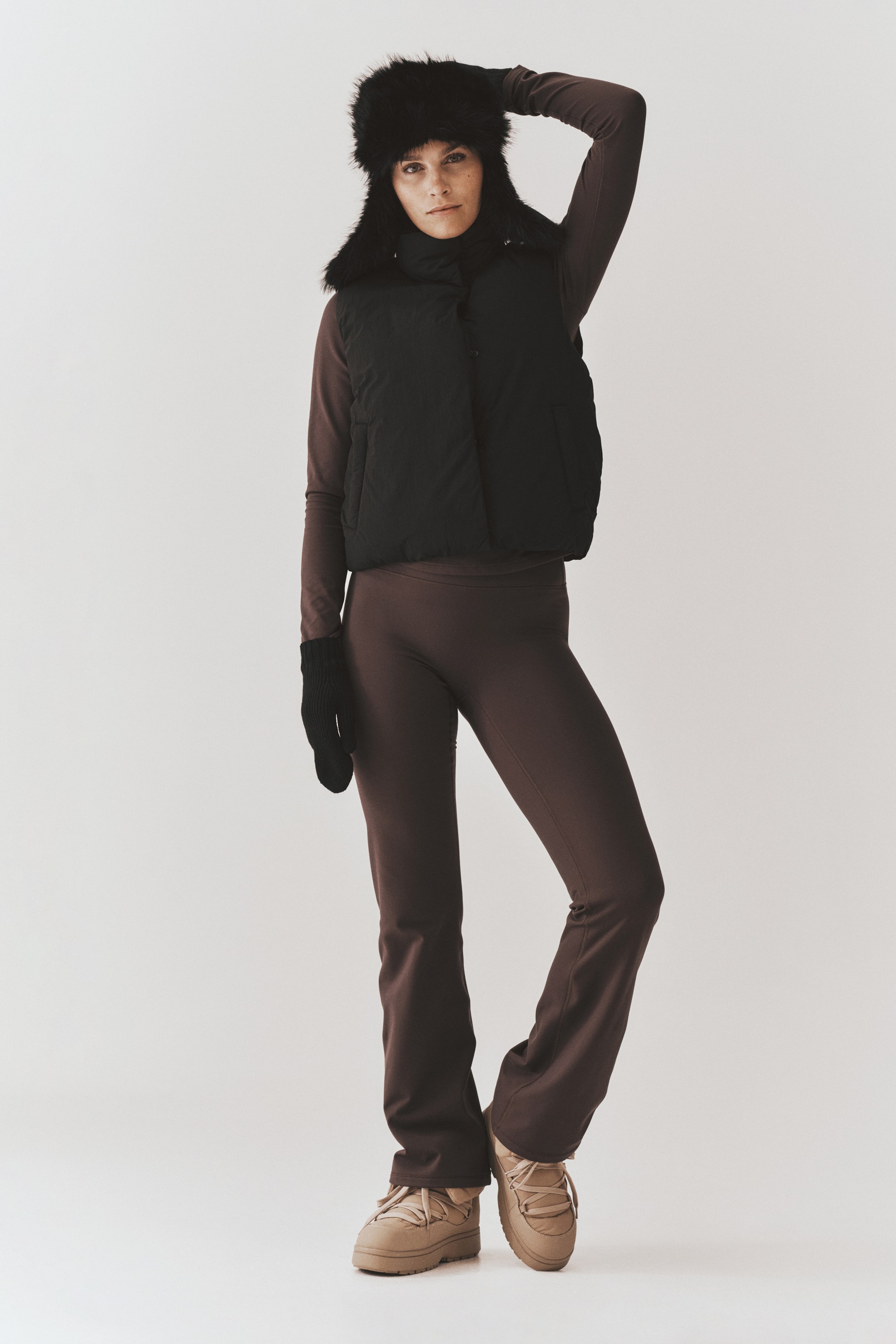 Größeres Bild ansehen: Ein schwarzer Steppgilet wird vom Model über einem braunen Langarmtop und einer dazu passenden ausgestellten Hose getragen. Das Outfit wird mit einer schwarzen Kunstfellmütze, schwarzen Strickfäustlingen und beigen Schnürstiefeln kombiniert.