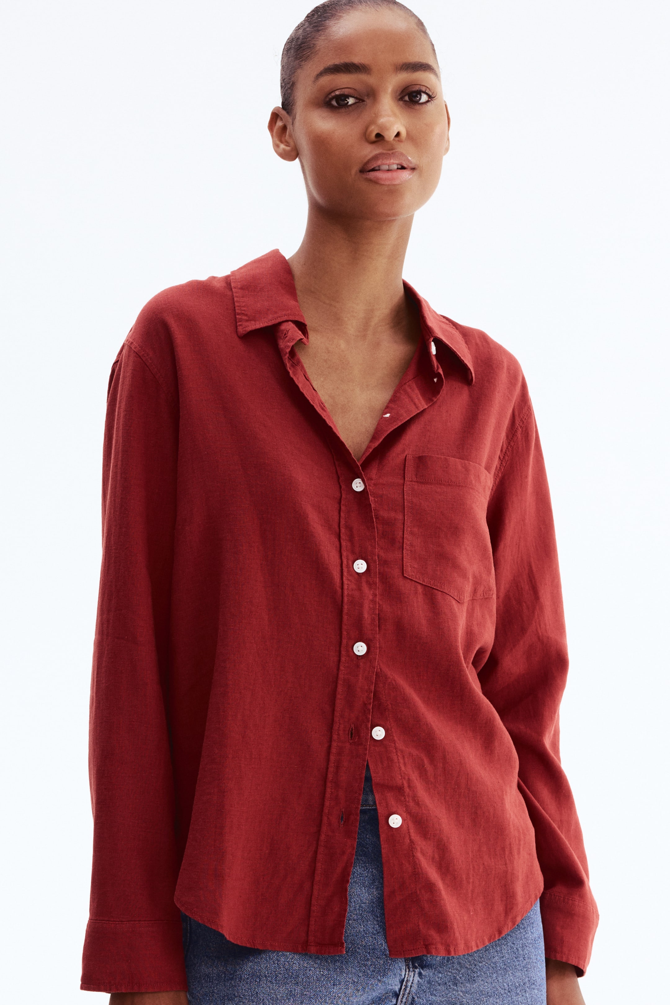 View larger image: A rust-red linen button-down shirt is presented front-facing, featuring a pointed collar and white buttons down the placket. It is tailored with long sleeves and a single chest pocket on the left, and is styled untucked over blue jeans.
