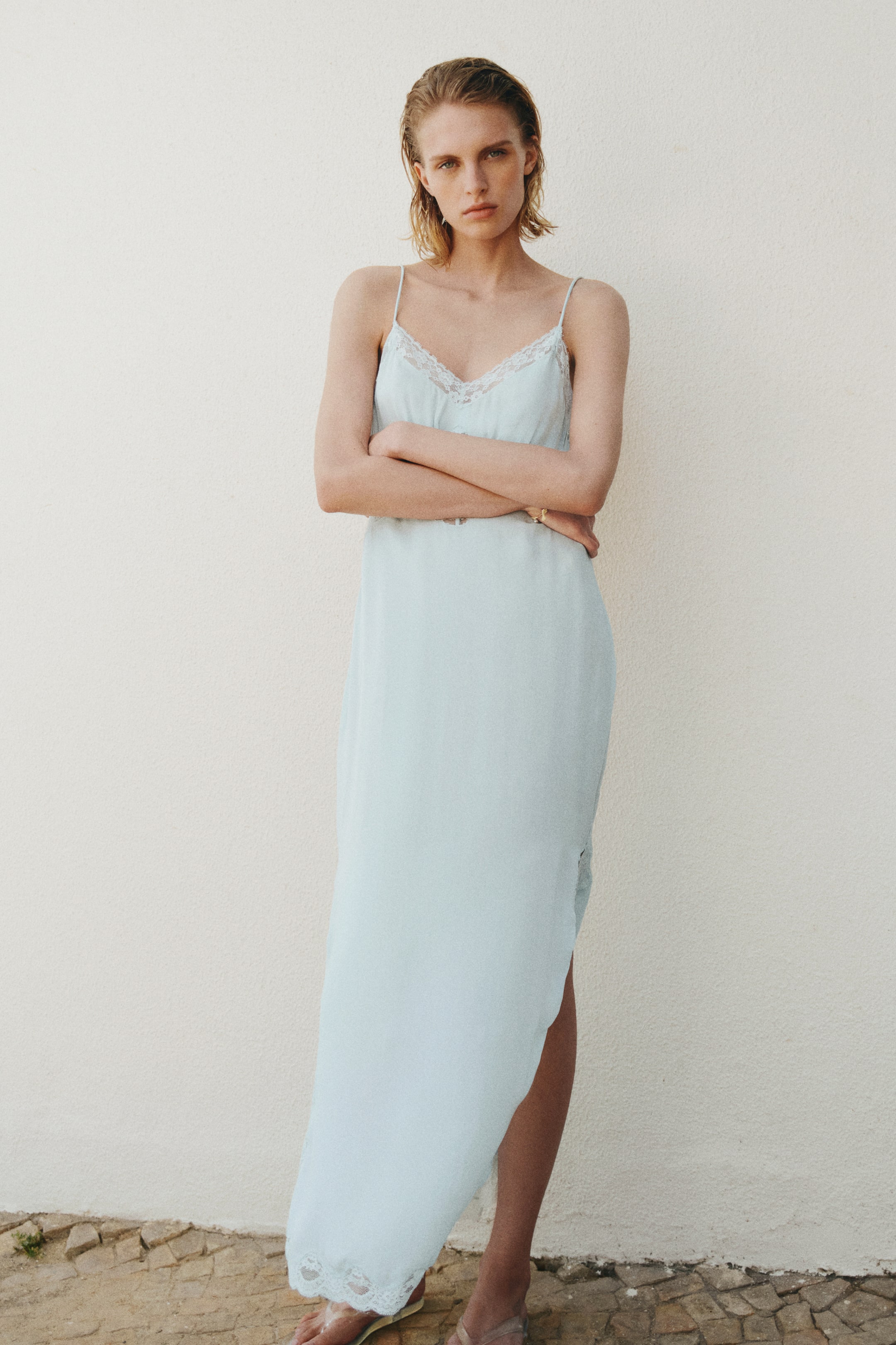 View larger image: The model is wearing a calf-length light blue dress with a V-neckline, adorned with scalloped lace trims and spaghetti shoulder straps. As she stands with her arms crossed, a high slit is noticeable at the side of the hem.