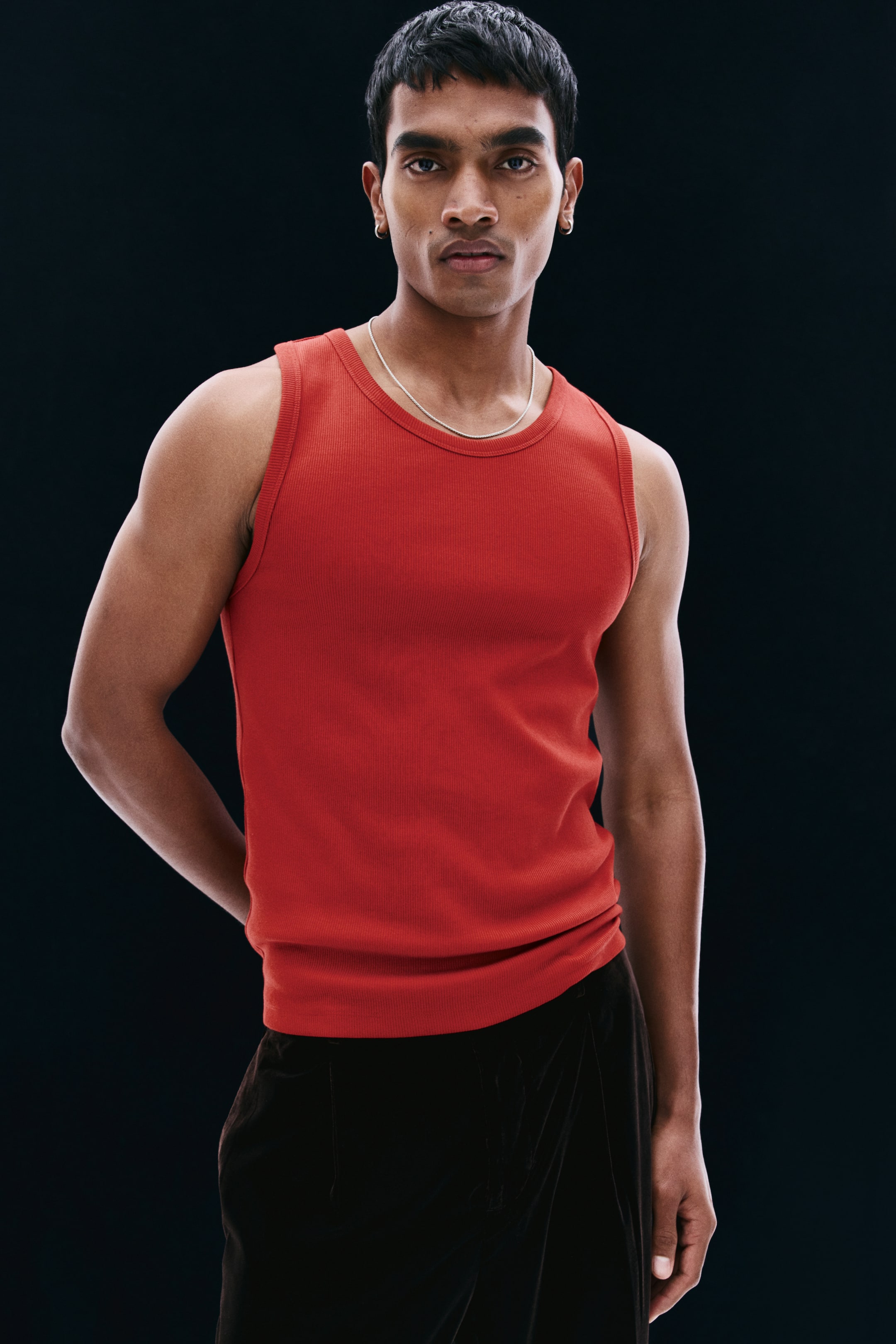 View larger image: A red ribbed cotton jersey tank top is being worn by the model, showcasing its slim fit, round neckline, and sleeveless design. The tank top is styled with dark trousers and a thin silver chain necklace.