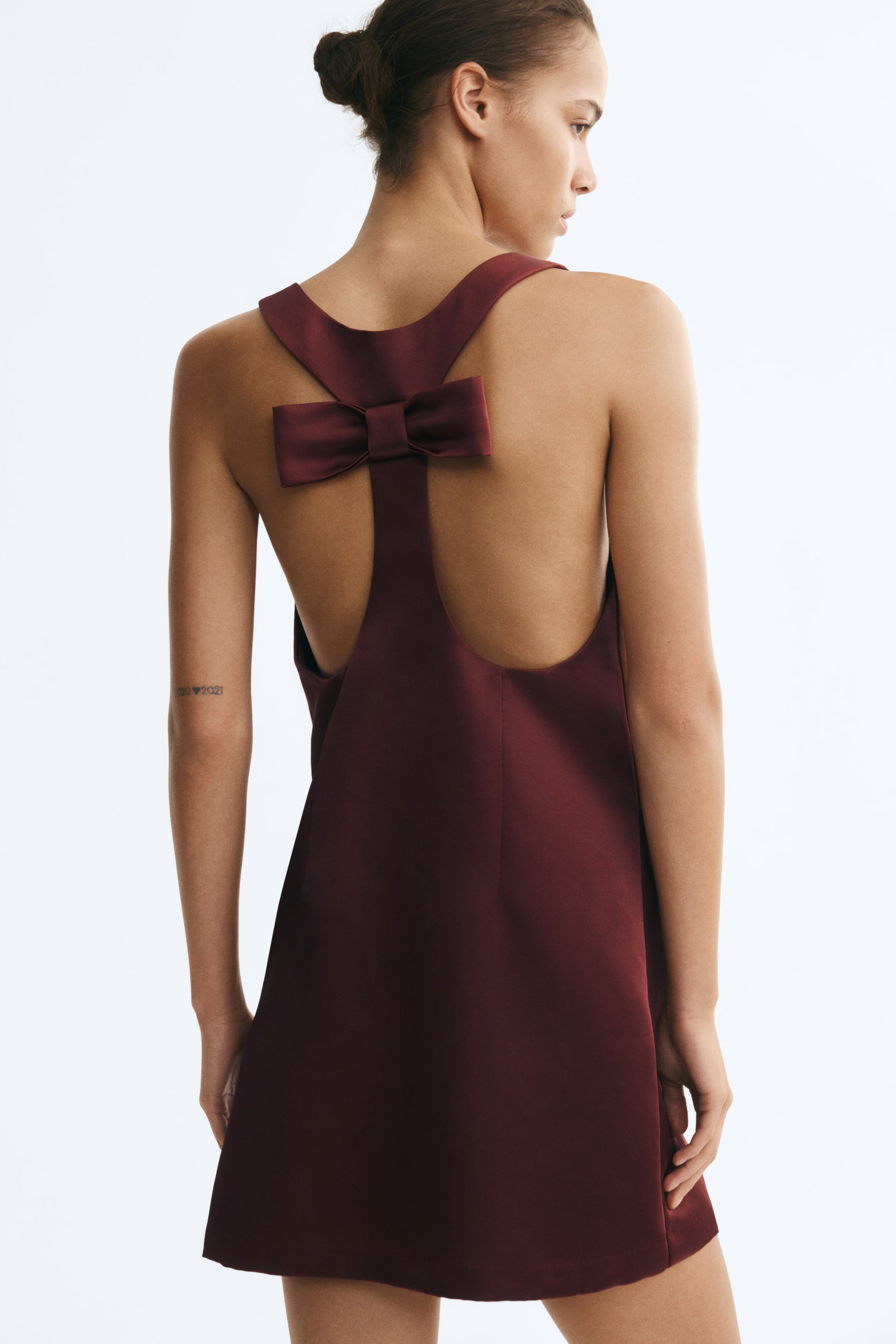 Visualizza immagine più grande: L'indossatrice indossa un abito corto in satin pesante bordeaux, mettendo in risalto il design del retro. L'abito presenta una schiena a vogatore impreziosita da un fiocco decorativo strutturato e profondi cut-out laterali.