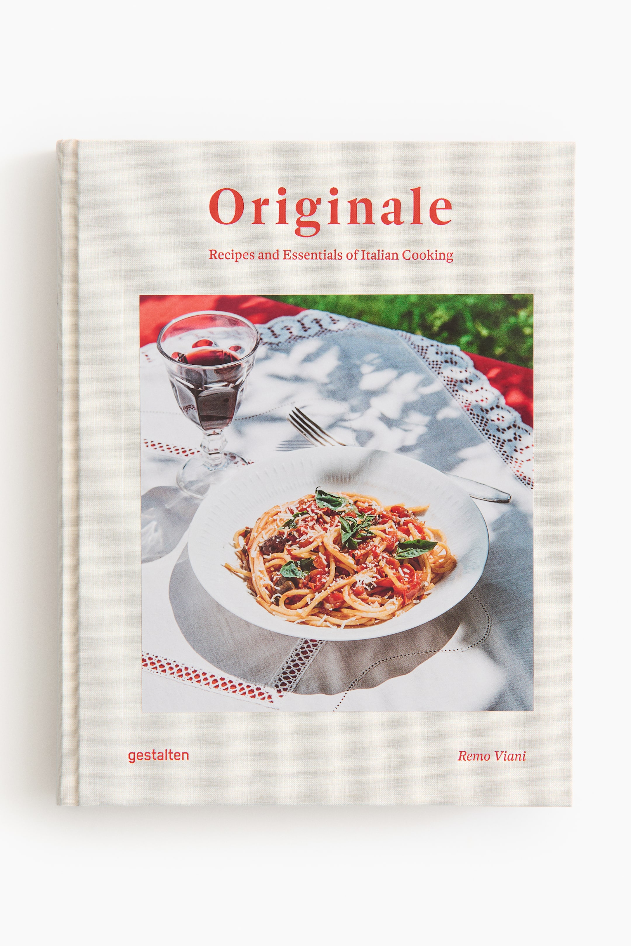 Vis større bilde: Boken viser sitt forsideomslag, laget av et lyse farget, teksturert stoff, med tittelen "Originale" og undertittelen "Oppskrifter og essensielle elementer i italiensk matlaging" i rød tekst. Et stort sentralt fotografi viser en tallerken med pasta med rød saus, ost og basilikum, ved siden av et glass rødvin på en mønstret duk, med "gestalten" og "Remo Viani" i rød tekst nederst.