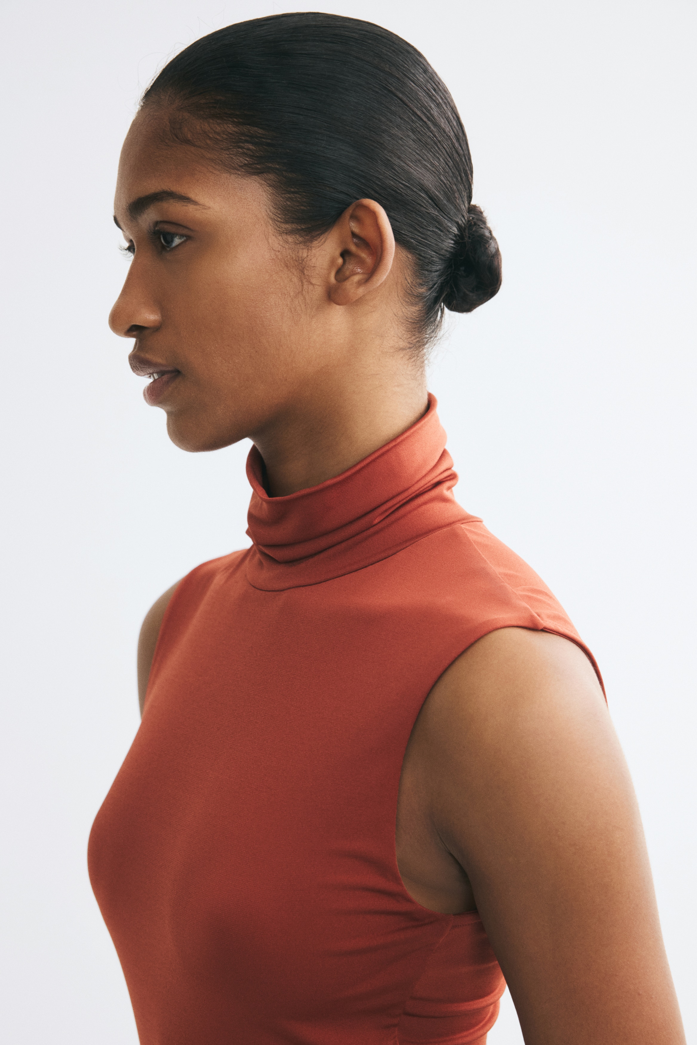 A rust-red sleeveless bodysuit is shown from the front, showcasing a draped roll neck and a fitted silhouette. The garment features a high-leg cut with a popper fastening at the bottom.