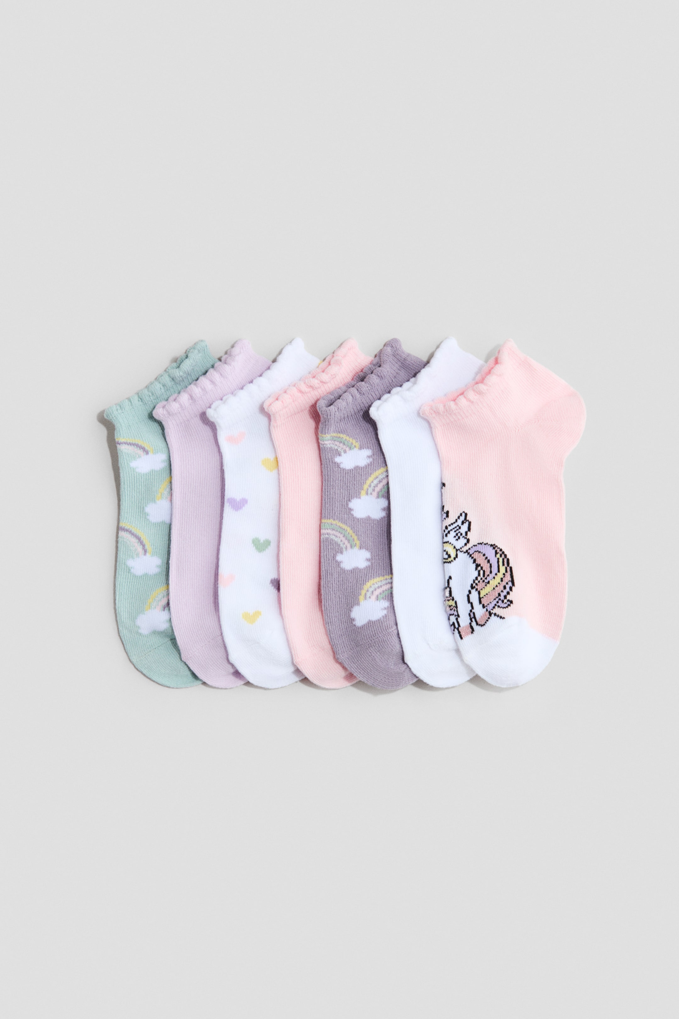 View larger image: Fine-knit trainer socks showcase a delightful array of pastel hues and patterns, adorned with ruffled elasticated tops. A set of seven unique socks is neatly lined up, each boasting charming designs like rainbows, hearts, and a unicorn.