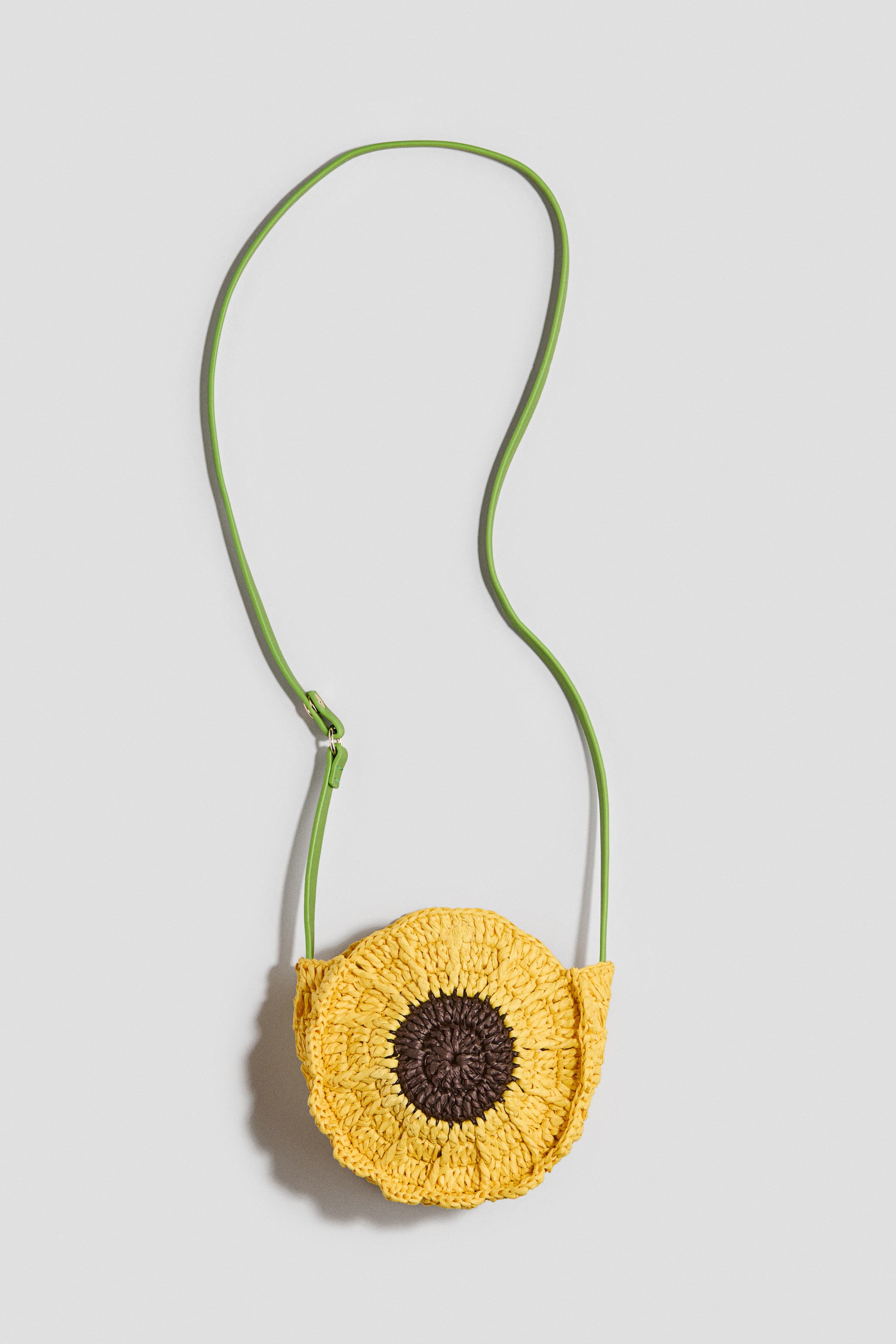 View larger image: A sunflower-shaped woven bag is displaying its front. It features yellow woven petals surrounding a dark brown woven centre. A thin lime-green strap extends from the top sides of the bag.