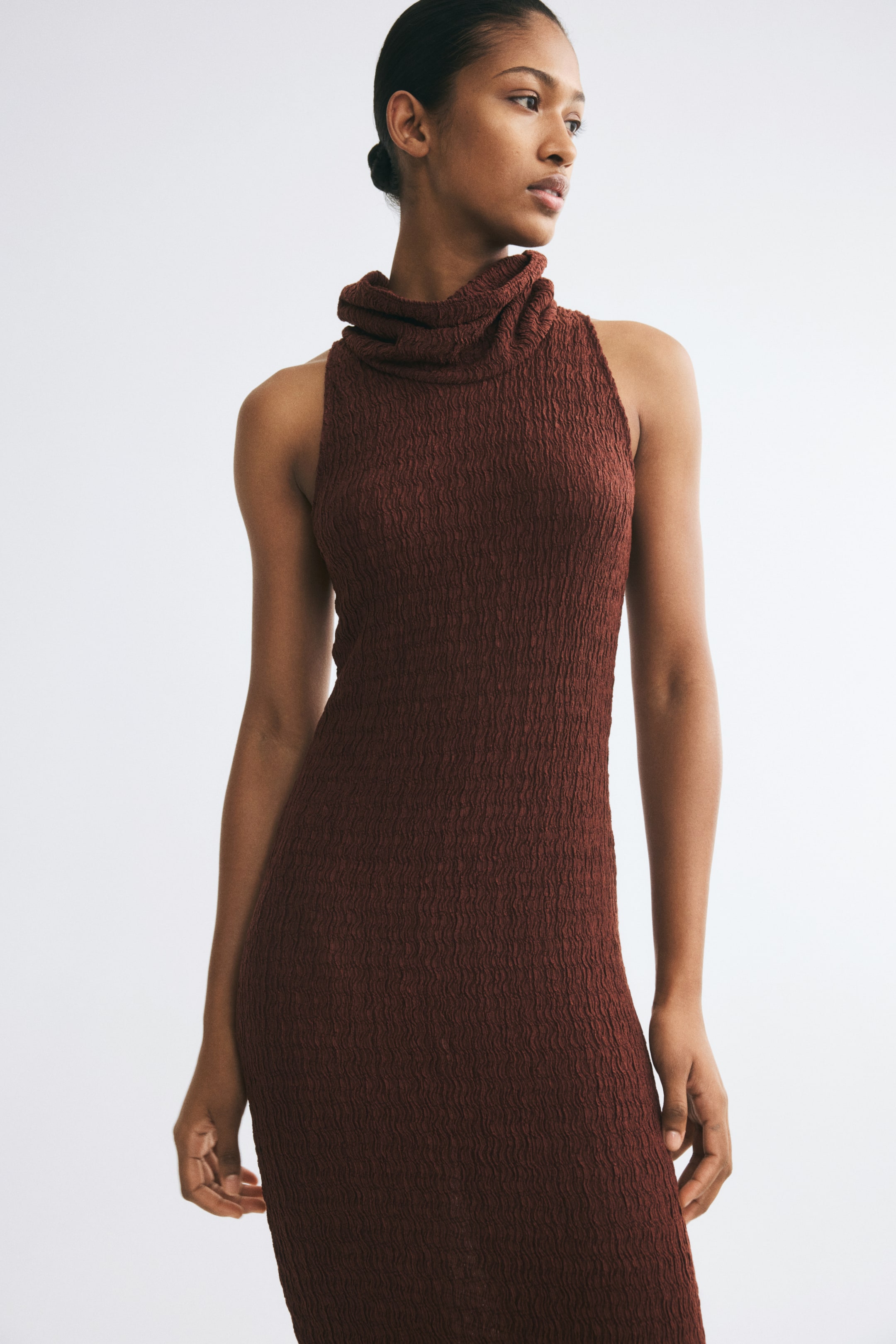 Ampliar la imagen: La modelo lleva puesto un vestido sin mangas, de corte ajustado, en jersey arrugado color café. El vestido presenta un escote drapeado tipo cowl con doblez, texturizado y muy llamativo, además de una silueta ceñida.