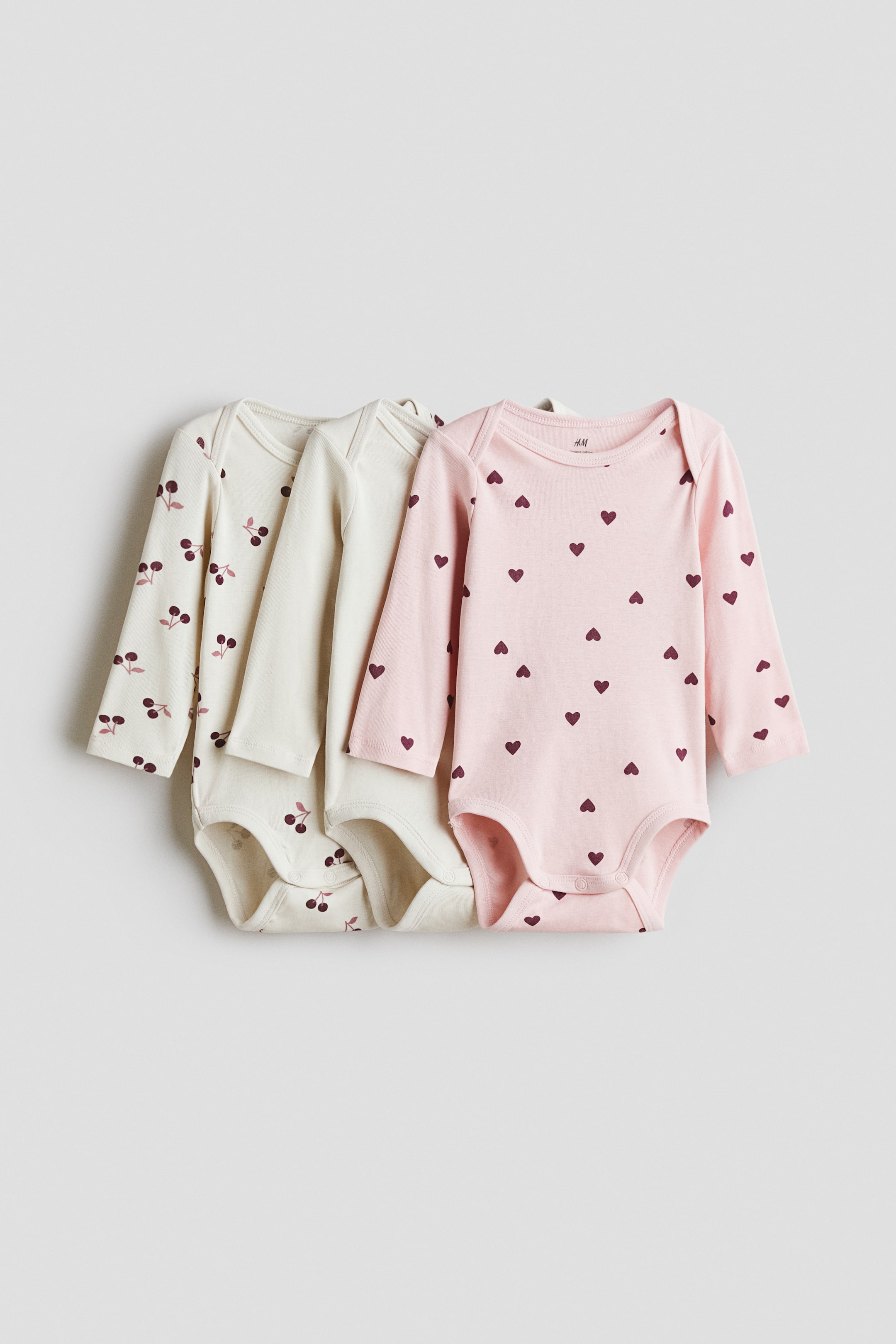 Grotere afbeelding bekijken: Drie babyrompers met lange mouwen worden van voren weergegeven, in een overlappende opstelling. De set bestaat uit een gebroken witte romper met kersenprint, een effen gebroken witte romper en een lichtroze romper met hartjesprint, allemaal met envelopehals en drukknoopsluitingen aan de onderkant.