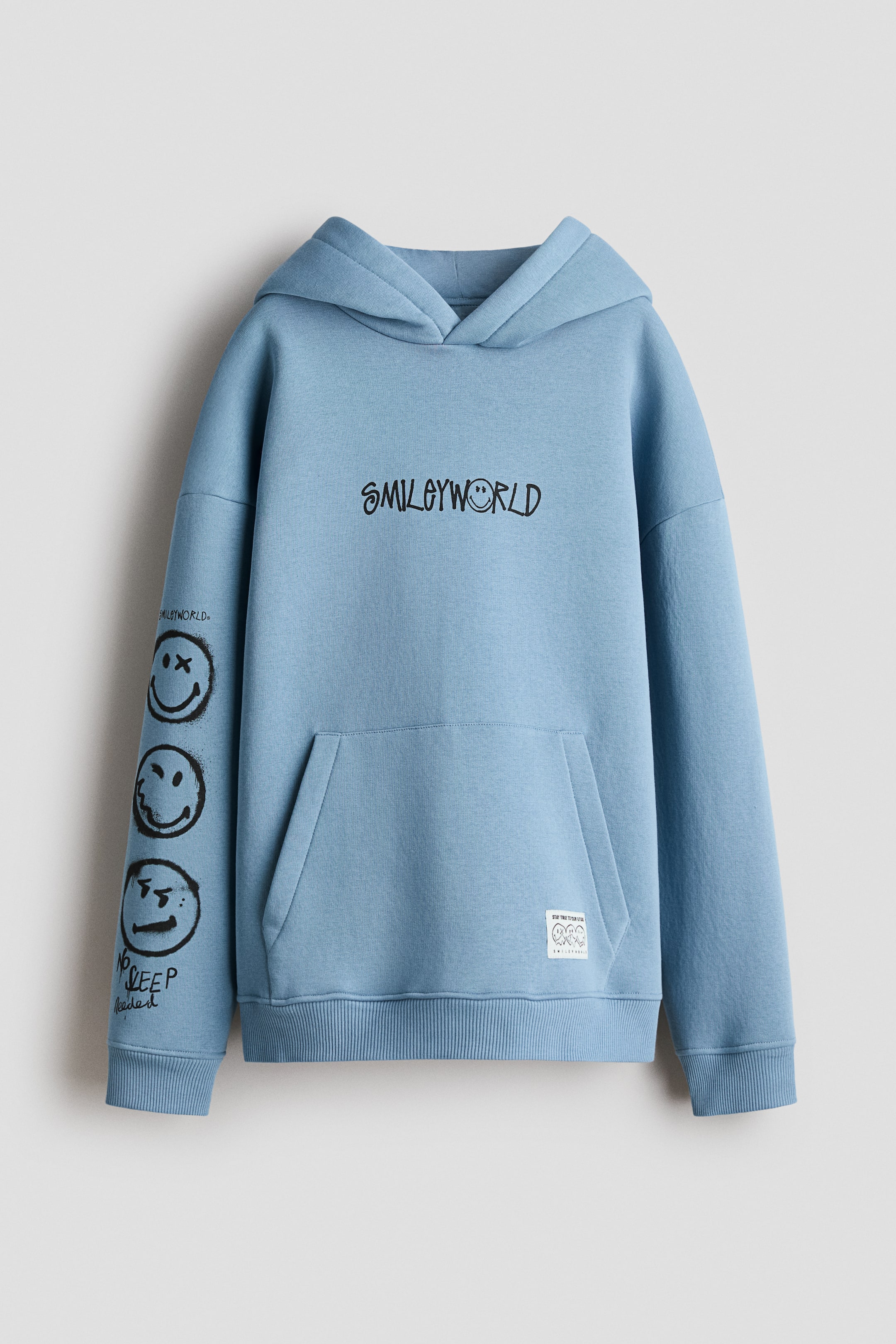 Visa större bild: En löst sittande ljusblå huvtröja i sweatshirt-tyg med tryck visar en "SMILEYWORLD"-grafik på bröstet och en serie smiley-ansikten med text på vänster ärm. Den har en fodrad huva med omlottfront, nedhasade axlar, en känguruficka med en liten vit märkesetikett samt ribbade muddar och fåll.