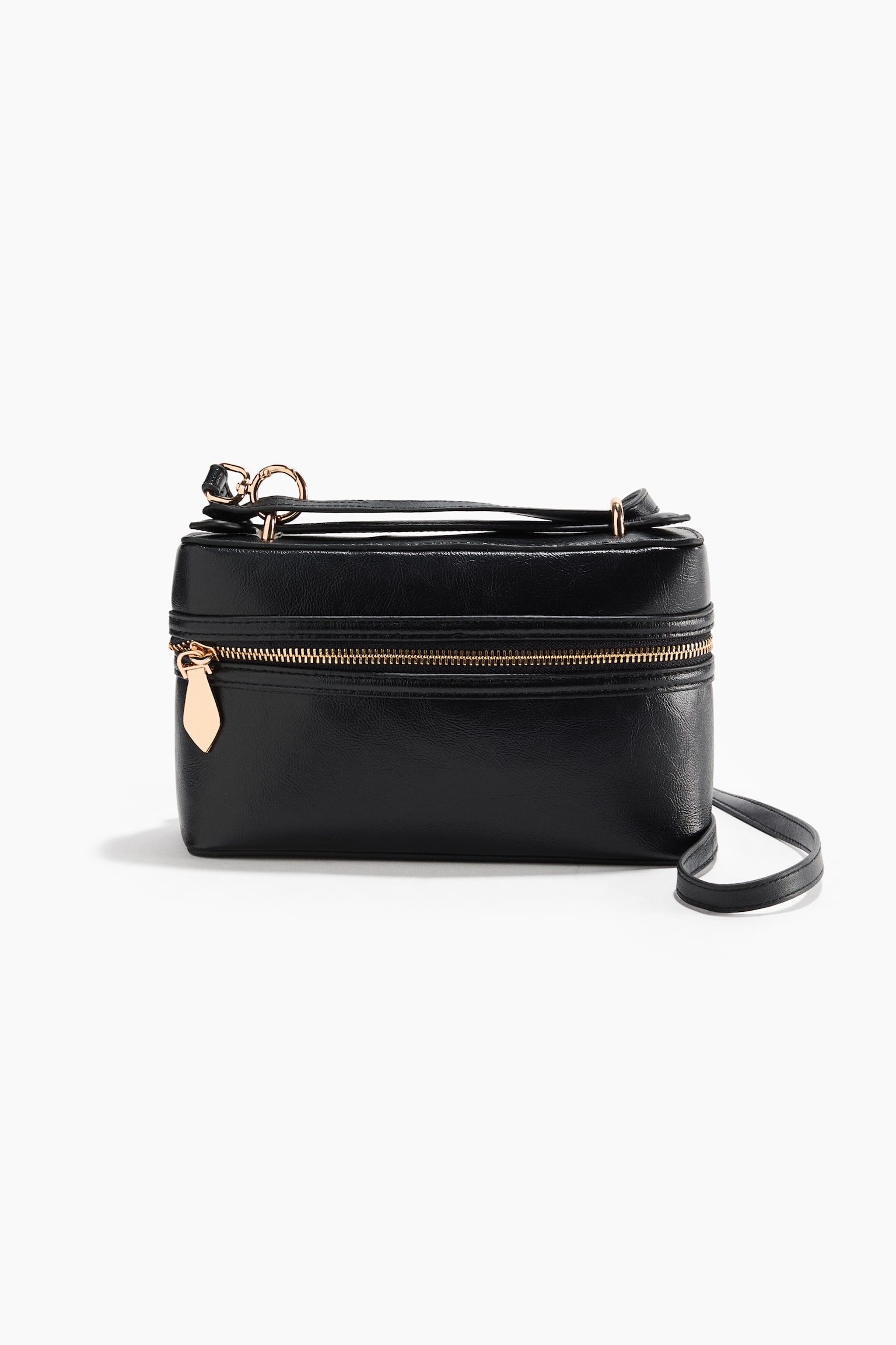 A black structured leather handbag is shown front-facing, showcasing its rectangular box shape with a horizontal gold-toned zip and a teardrop-shaped zip pull. A flat top handle lies across the top, secured by gold-toned metal rings, while a slim black shoulder strap extends from the right side.