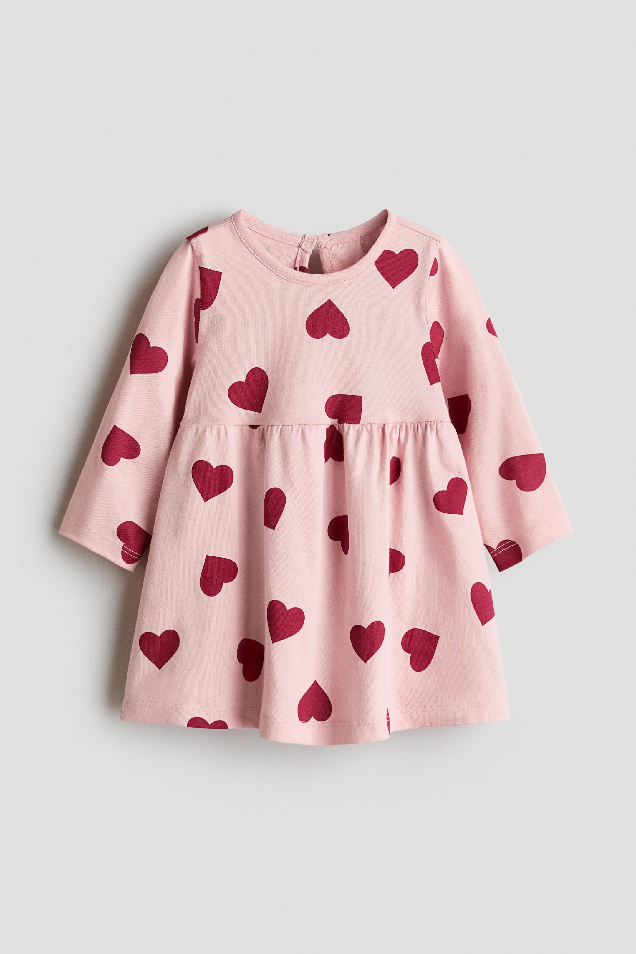 View larger image: A pink baby dress featuring a scattered red heart pattern. Designed with long sleeves, a round neckline, a keyhole button closure at the back of the neck, and a gathered waist that creates a flared skirt.