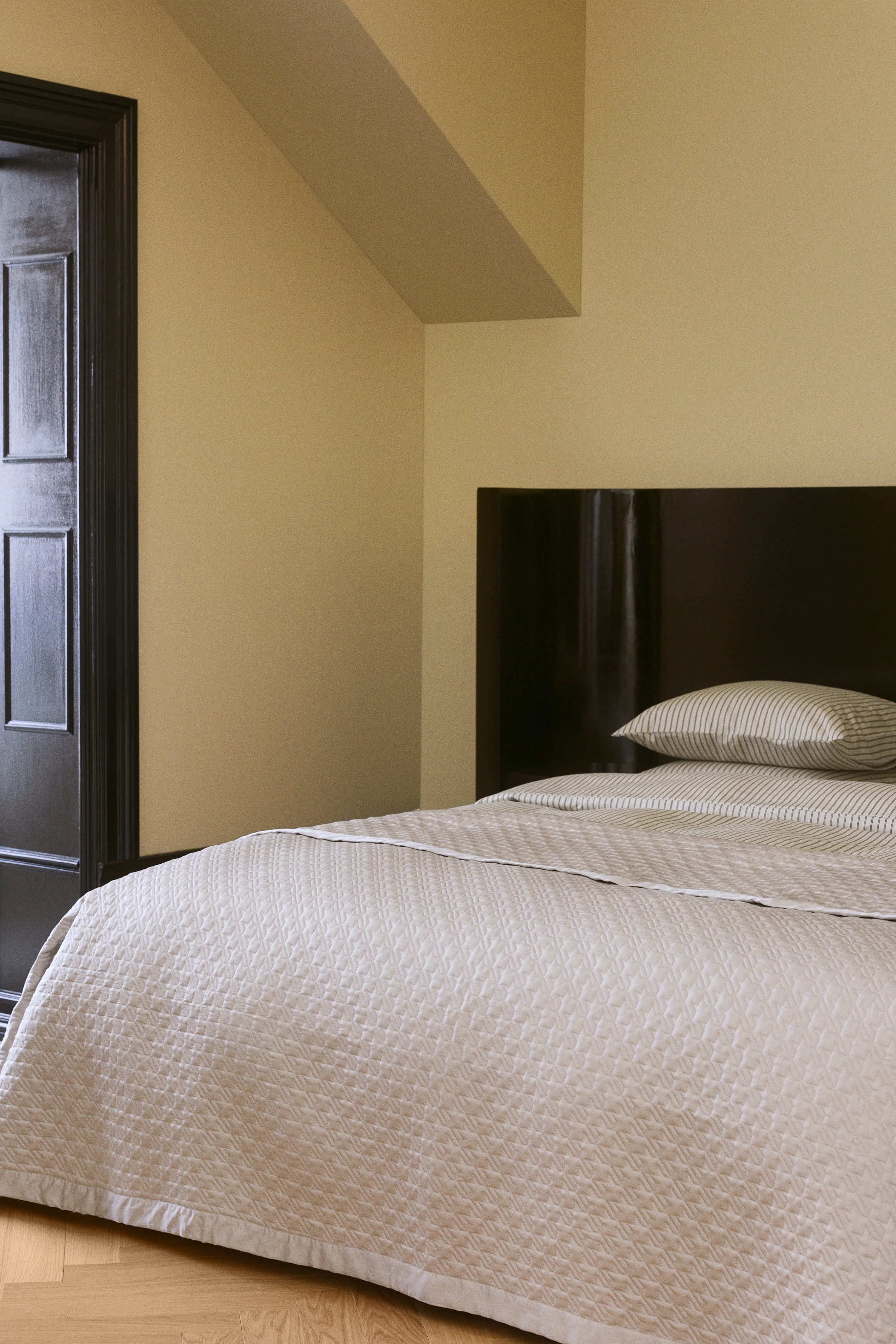 A light beige blanket is lying flat, featuring a quilted surface with a repeating houndstooth pattern. A smooth, unquilted border is framing all four edges of the blanket.