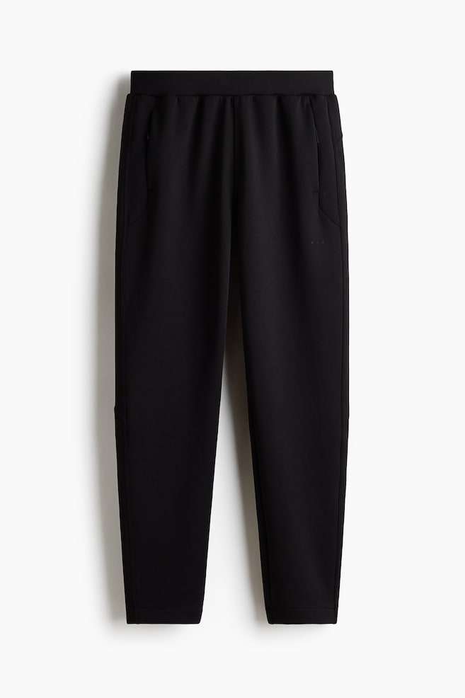 Los pantalones negros presentan una pretina elástica y bolsillos laterales, uno de los cuales cuenta con un cierre de cremallera en el lado izquierdo. Un sutil texto "DO R" aparece en el muslo derecho, y los dobladillos de los pantalones son lisos. - 1