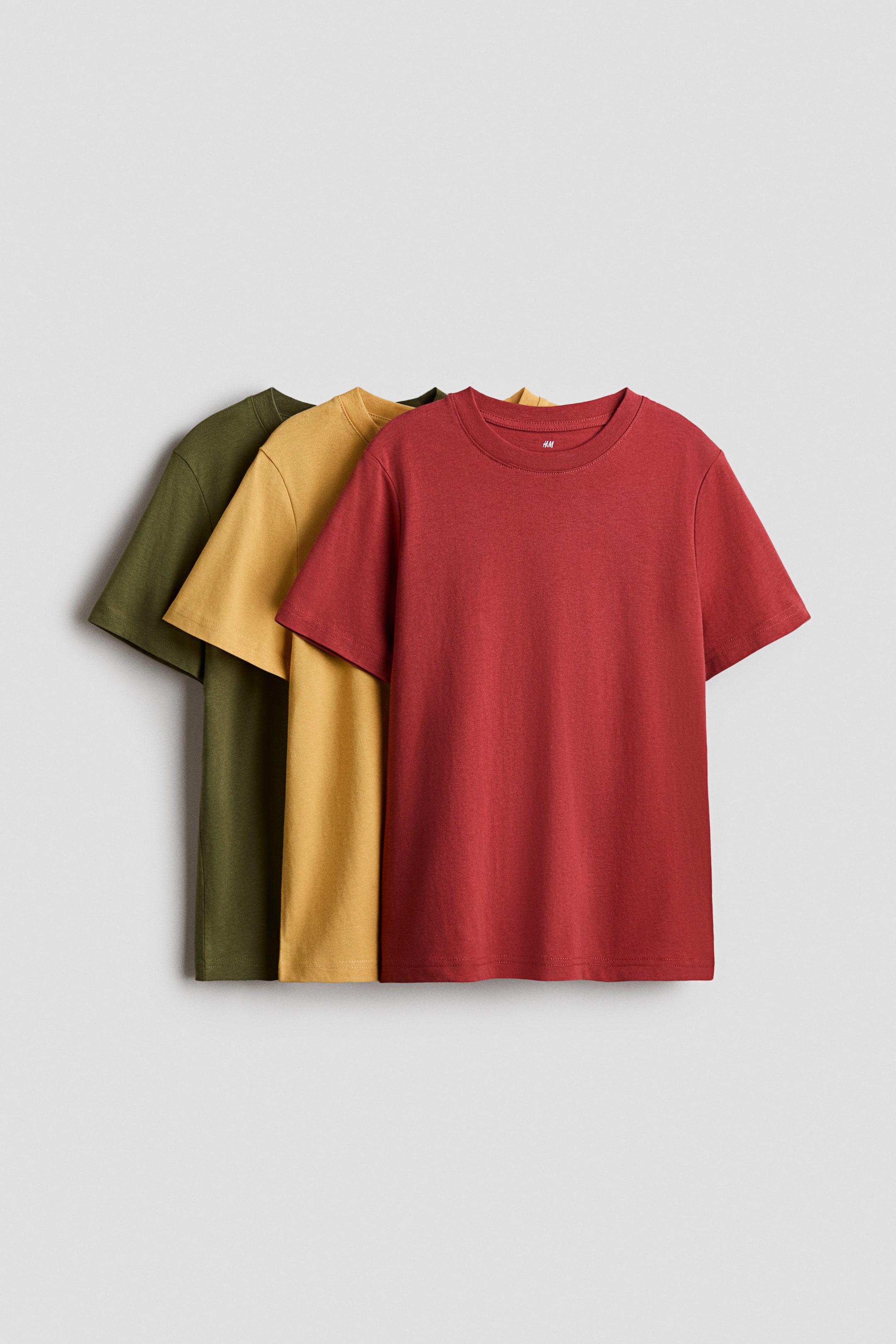 View larger image: "Round-necked T-shirts crafted from soft cotton jersey are showcased as a stack of three. At the forefront, a short-sleeved red T-shirt is prominently displayed, with a small white tag visible inside its neckline. Behind it, the short-sleeved mustard yellow and olive green T-shirts are elegantly layered."
