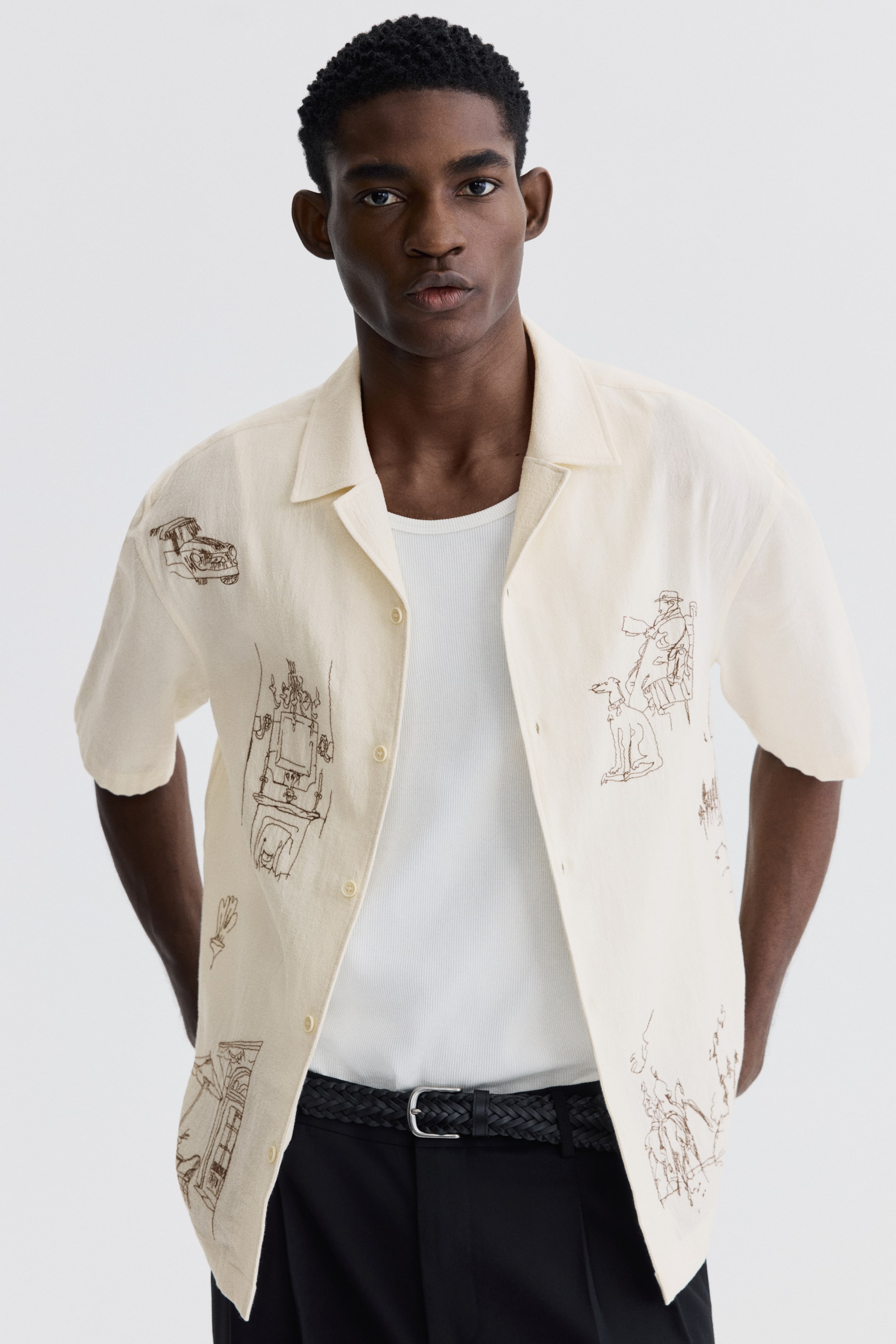 查看大圖: 這款奶油色短袖襯衫採用露營領設計，表面上有精緻的棕色線條圖案。襯衫敞開穿在白色羅紋背心外，展示出其前扣設計和未紮的下擺。