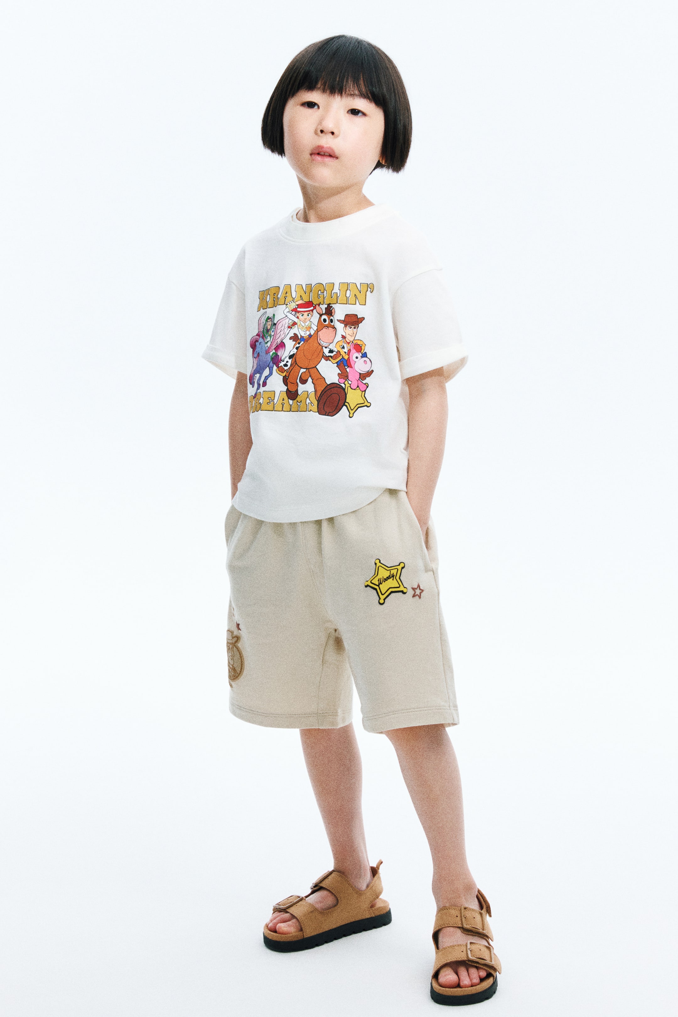 拡大画像を表示: 前面から紹介されているのは、半袖のオフホワイトのTシャツとベージュのショーツです。Tシャツには、カラフルな漫画キャラクターのグラフィックと「Wranglin' Team」というテキストが胸に描かれており、袖口がカフスになっています。ベージュのショーツは、ウエストがゴム仕様で、サイドポケットがあり、左足には「Woody」と刺繍された黄色の保安官の星のパッチが、右足にはブラウンのカウボーイハットが刺繍されており、膝上までの長さです。
