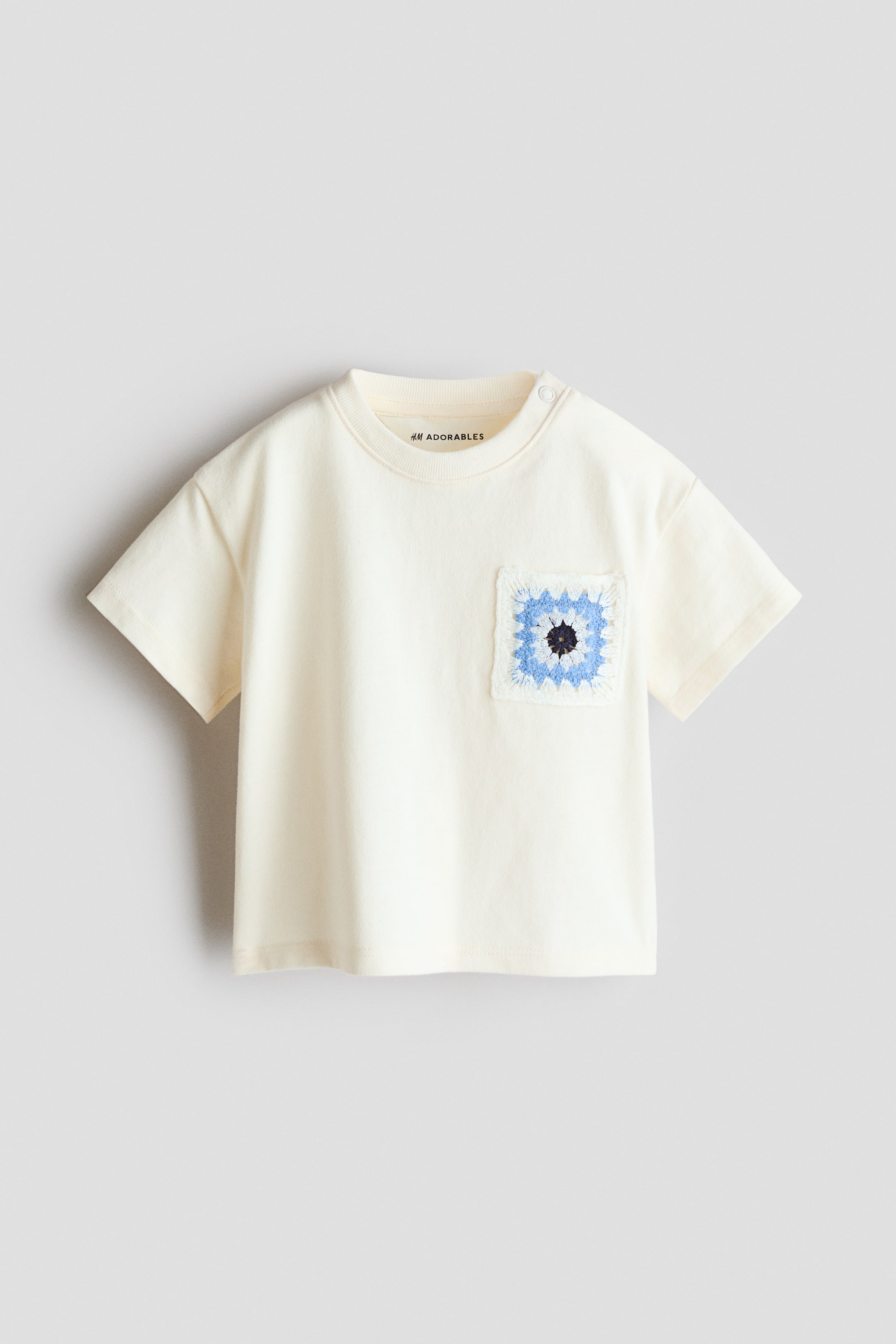 Grotere afbeelding bekijken: Losvallend T-shirt. Dit crèmekleurige T-shirt heeft korte mouwen en een geribde ronde hals. Een vierkante gehaakte borstzak, met een blauw, wit en zwart patroon en licht rafelige randen, siert de linkerborst, en een enkele drukknoop is zichtbaar op de linkerschouder.