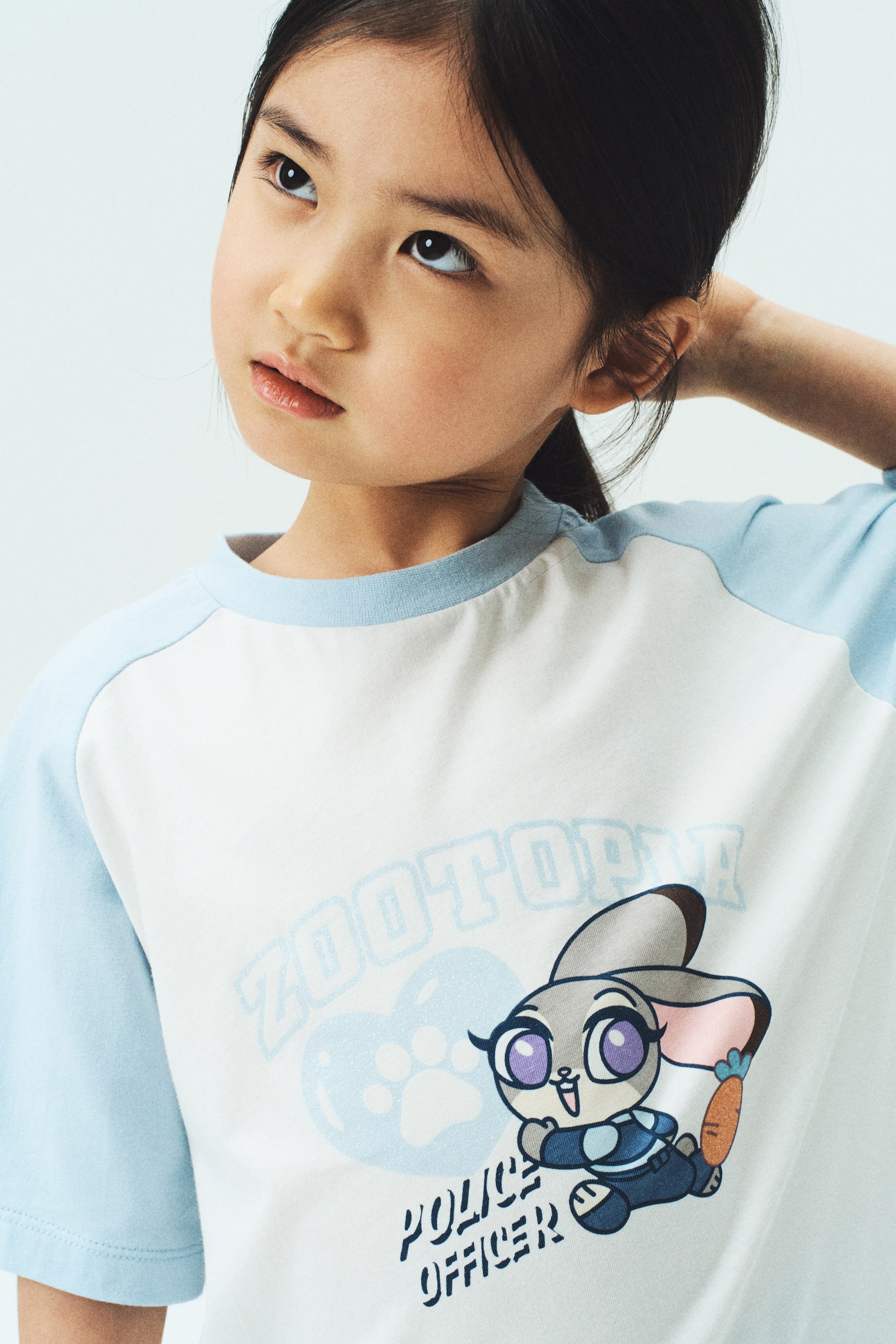 拡大画像を表示: 白い半袖ラグランTシャツが紹介されています。ライトブルーのラグランスリーブとリブ付きのライトブルークルーネックを特徴としています。フロントには、警察の制服を着た漫画のウサギキャラクターのグラフィックプリントがあり、キャラクターの上には「ZOOTOPIA」のテキストと肉球ハートデザインが、下には「POLICE OFFICER」のテキストがプリントされています。