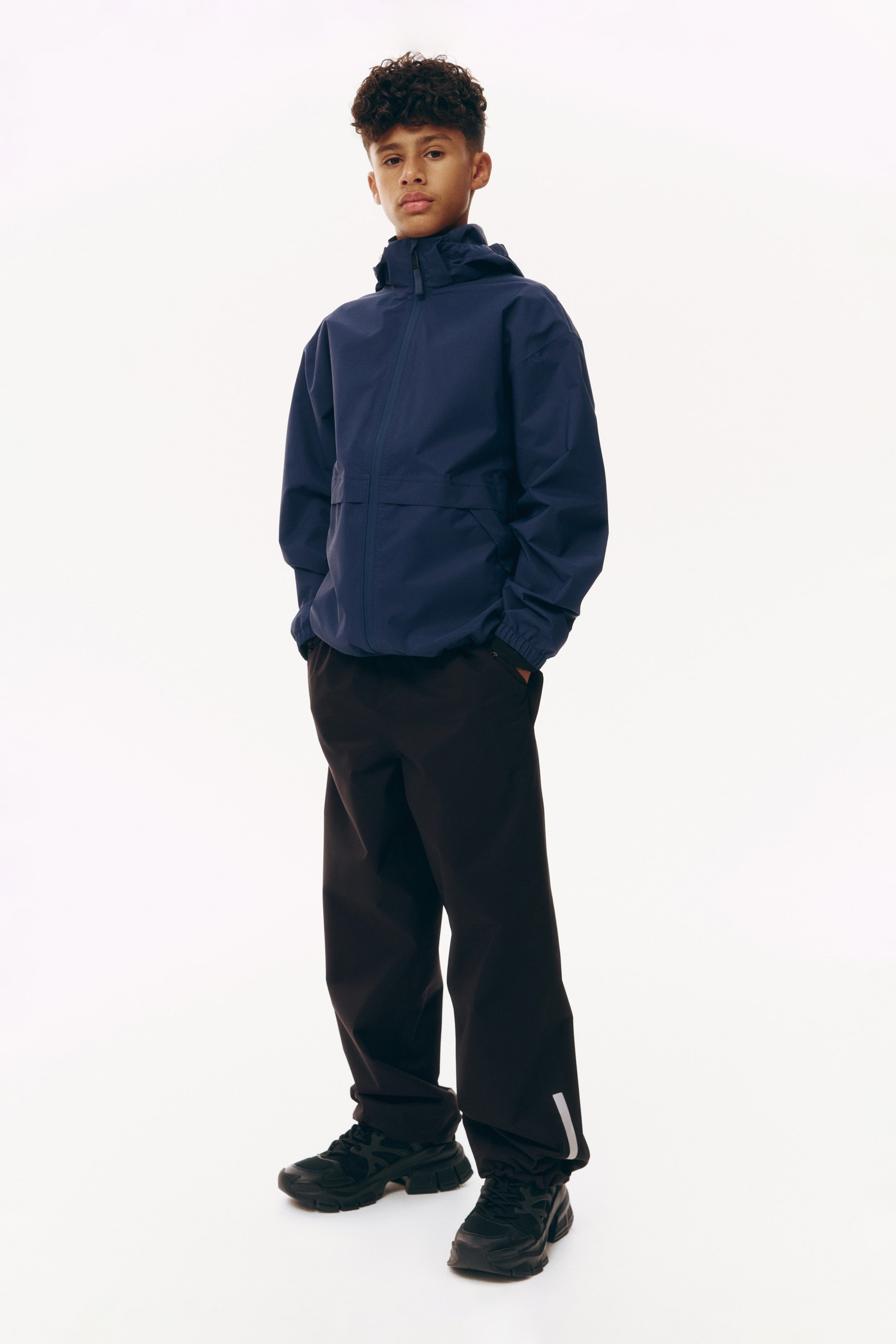 Vezi imaginea mai mare: O jachetă cu glugă de culoare albastru marin este prezentată frontal, având un fermoar complet, un guler înalt, manșete elastice și o cusătură orizontală pe piept. Este stilizată cu pantaloni negri cu croială lejeră, care prezintă o bandă reflectorizantă pe partea inferioară a piciorului drept, și adidași negri cu talpă groasă.