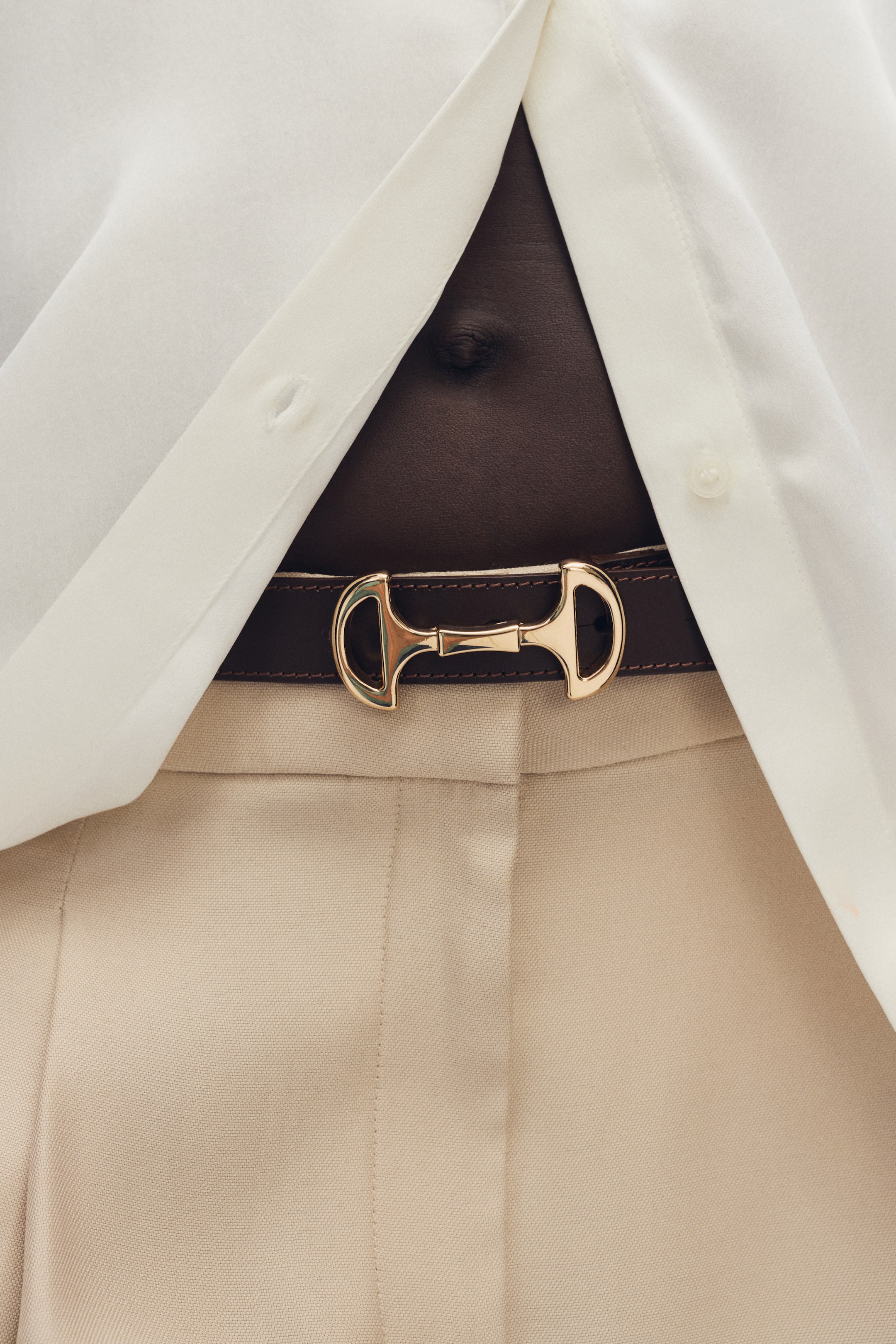 Visualizza immagine più grande: Una cintura in pelle marrone scuro con una fibbia a morsetto in metallo dorato è indossata intorno alla vita del modello, assicurando pantaloni beige chiaro. Una camicia bianca è parzialmente aperta, rivelando la cintura e il ventre del modello.