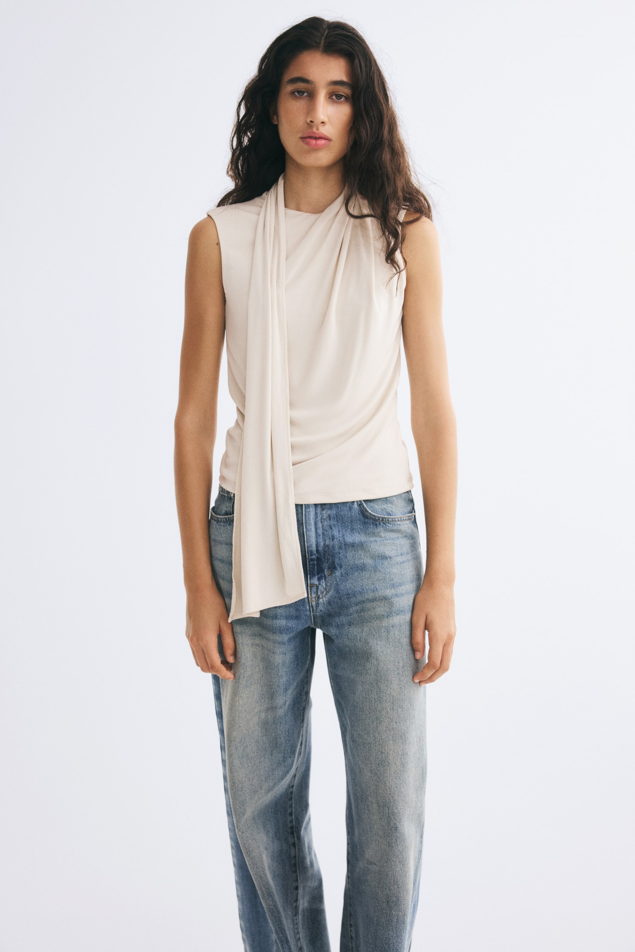 View larger image: The model is wearing a sleeveless light beige stretch jersey top with a draped scarf collar and straight hem, styled with blue denim jeans.