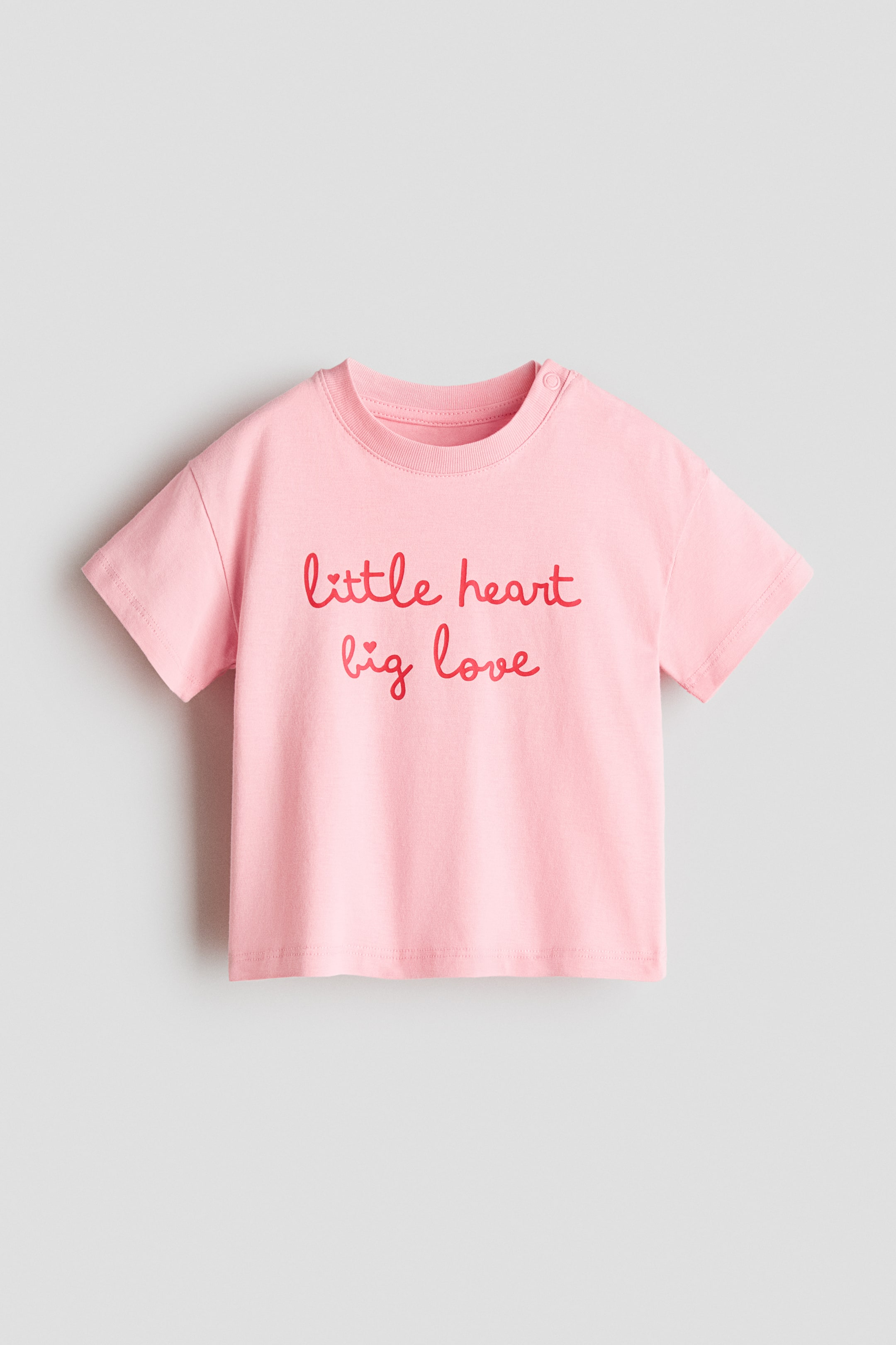 拡大画像を表示: ピンクのTシャツの前面には、胸に「little heart big love」と赤い文字が書かれています。リブ付きクルーネック、半袖、そして右肩に2つのスナップボタンがデザインされています。