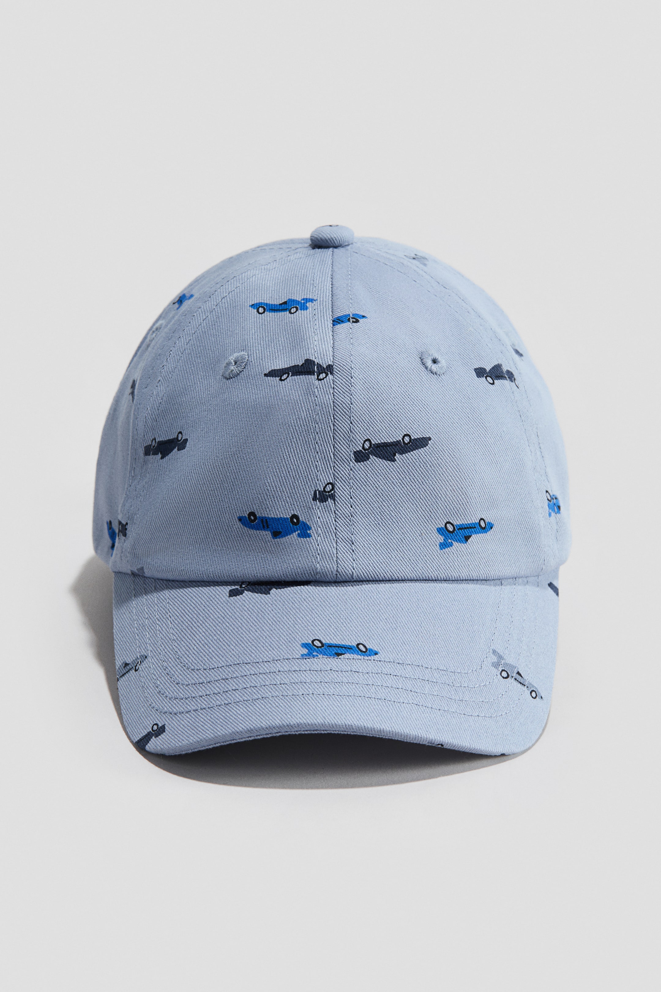 View larger image: A light blue cap is facing forward, featuring a woven fabric with a repeating pattern of small blue and dark gray race cars.
