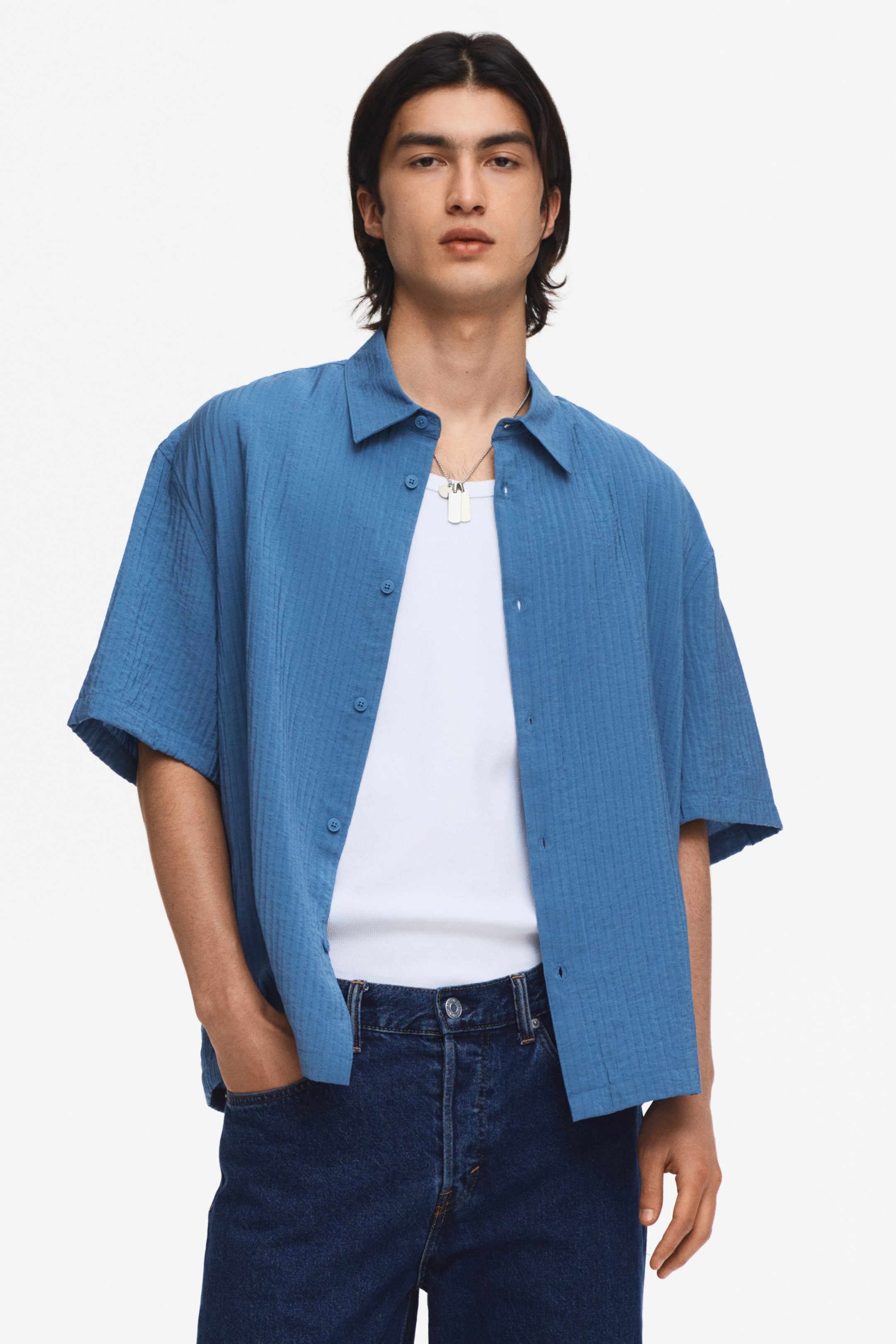 View larger image: A blue short-sleeved shirt, facing forward, is worn open over a white ribbed undershirt. It showcases a spread collar, visible dark buttons down the front placket, and a textured fabric with subtle vertical lines.
