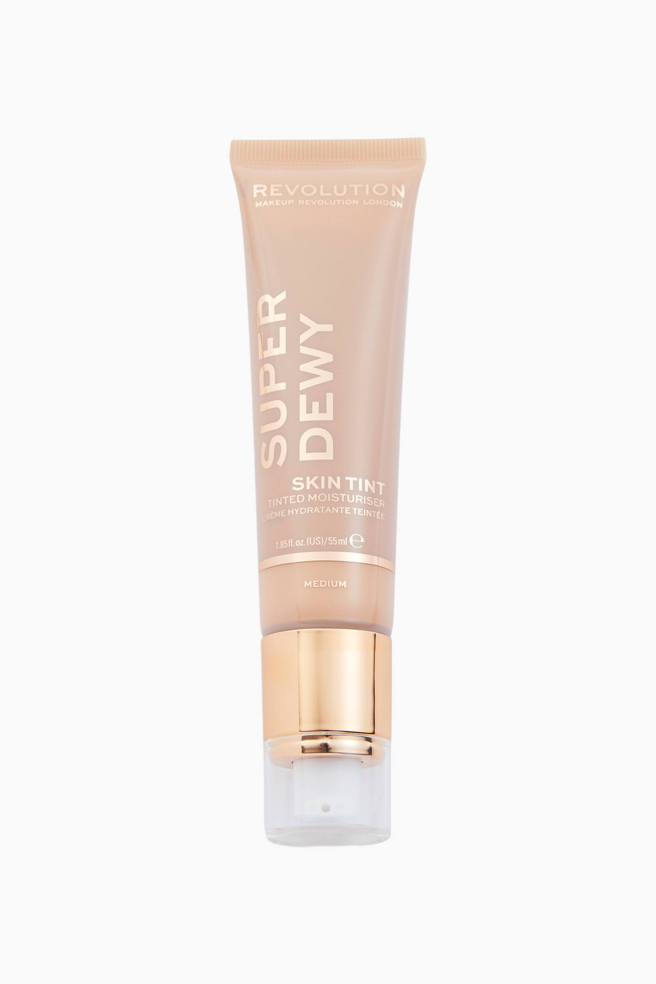 Visa större bild: Superdewy Tinted Moisturiser står upprätt i en framåtriktad vy och visar upp sin persikotonade klämförpackning som smalnar av till en pumpdispenser i metallicguld med en transparent plastkork. Tuben visar vit och guldtryckt text, inklusive ”REVOLUTION”, ”SUPER DEWY” och nyansnamnet ”MEDIUM” längst ned.