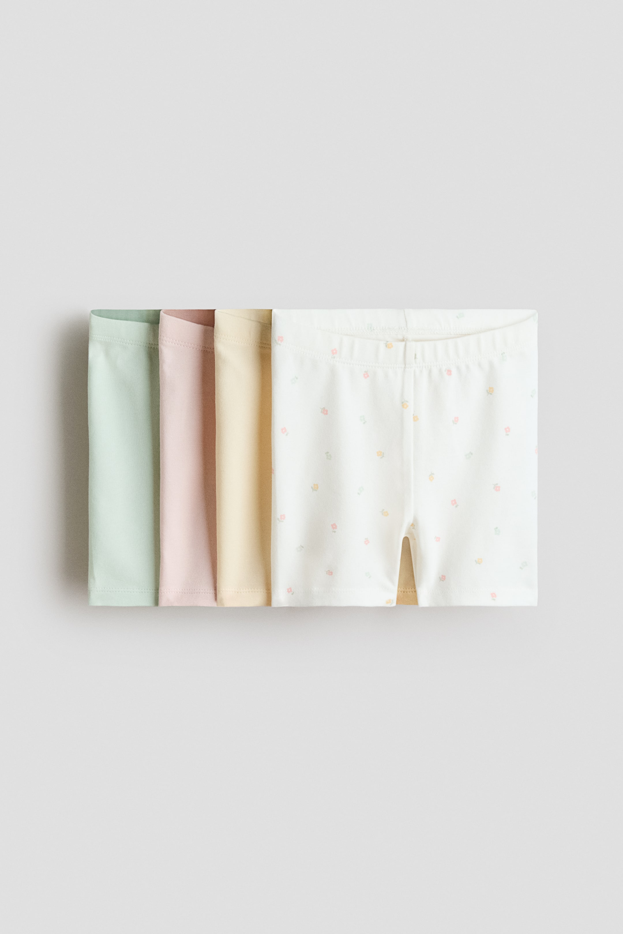 View larger image: Four pairs of cycling shorts are arranged horizontally, with a slight overlap. The front-most pair, in an off-white hue, features a scattered design of small, multicolored floral patterns and an elasticated waistband. Beneath it, three solid-colored pairs in light green, light pink, and light yellow are partially visible.