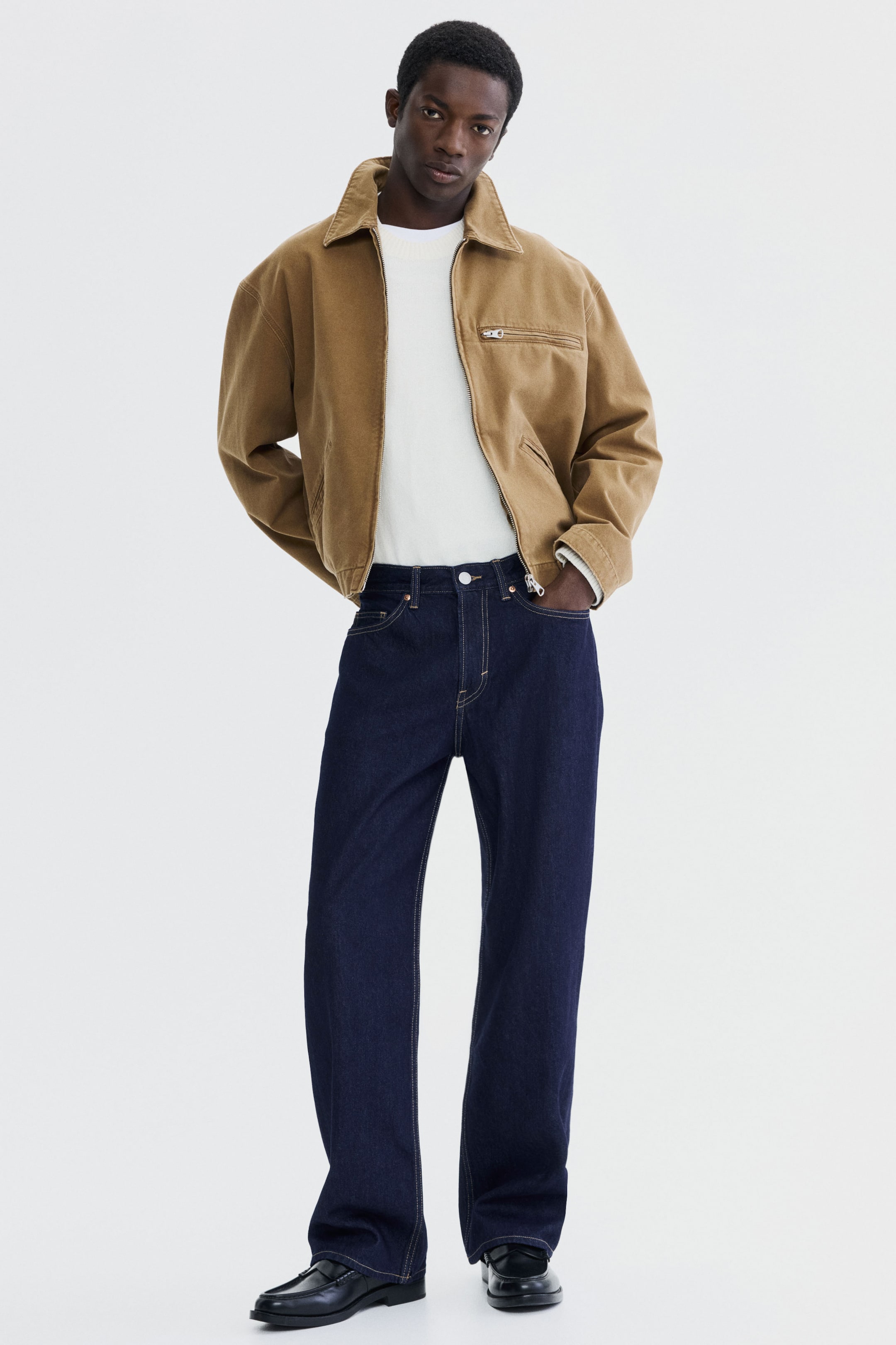 Xem ảnh lớn hơn: Áo khoác lao động màu nâu nổi bật với cổ bẻ, khóa kéo phía trước toàn bộ, túi ngực có khóa kéo bên trái và hai túi vá ở eo. Áo có kiểu dáng rộng rãi và được mặc bên ngoài áo thun trắng, kết hợp với quần jean xanh đậm.