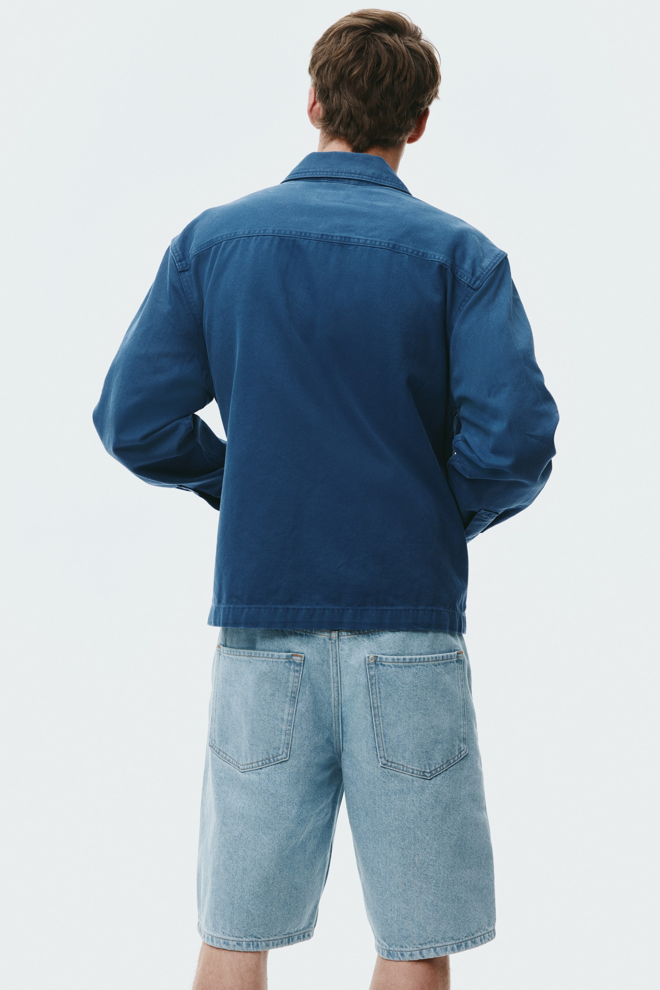 Größeres Bild ansehen: Eine blaue Jacke mit Kragen, langen Ärmeln und geradem Saum wird vom Modell getragen, das mit dem Rücken zur Kamera steht und die Hände in den Taschen der Jacke hat. Die Jacke liegt über hellgewaschenen Denim-Shorts mit Gesäßtaschen, die bis zum Knie reichen.