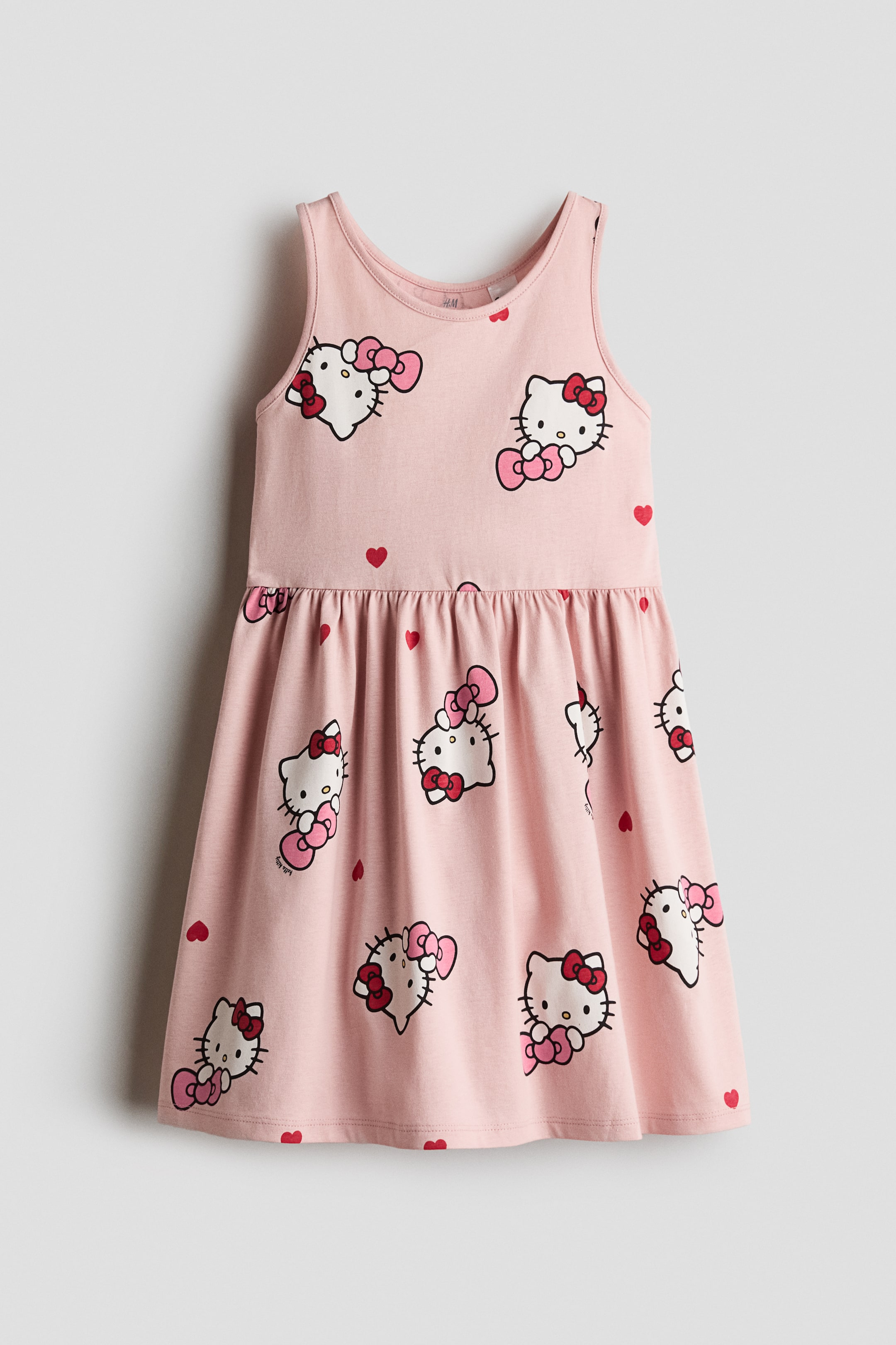View larger image: A pink, sleeveless cotton jersey dress is shown from the front, featuring a round neckline, a gathered seam at the waist, and a flared skirt with an all-over print of Hello Kitty faces and small red hearts.