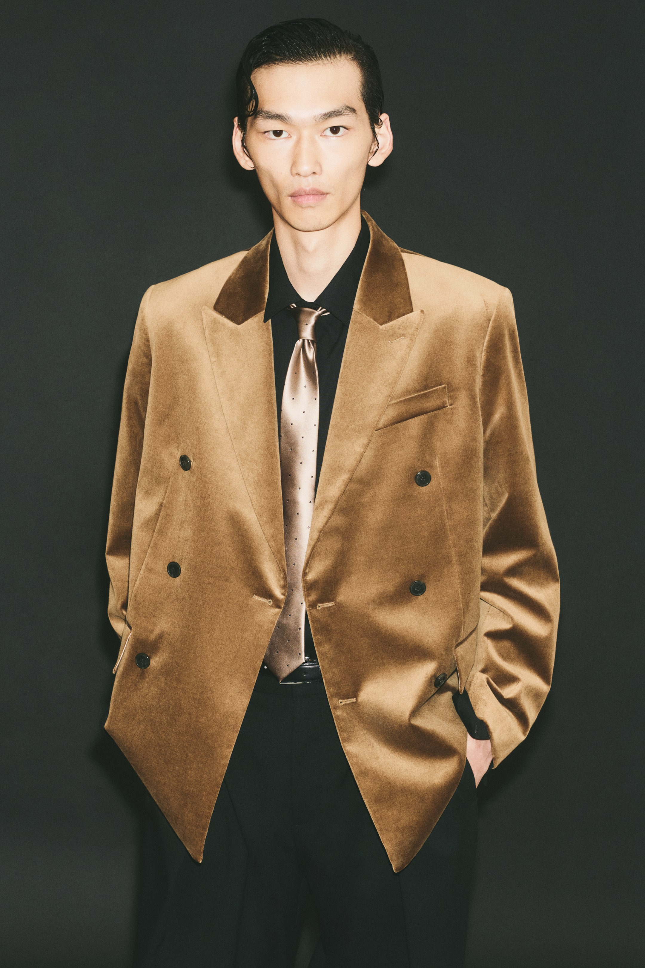 View larger image: A golden-brown double-breasted jacket featuring peak lapels, a welt chest pocket, and dark buttons is being worn open by the model. The jacket is layered over a black button-down shirt, a bronze-toned patterned tie, and black trousers.