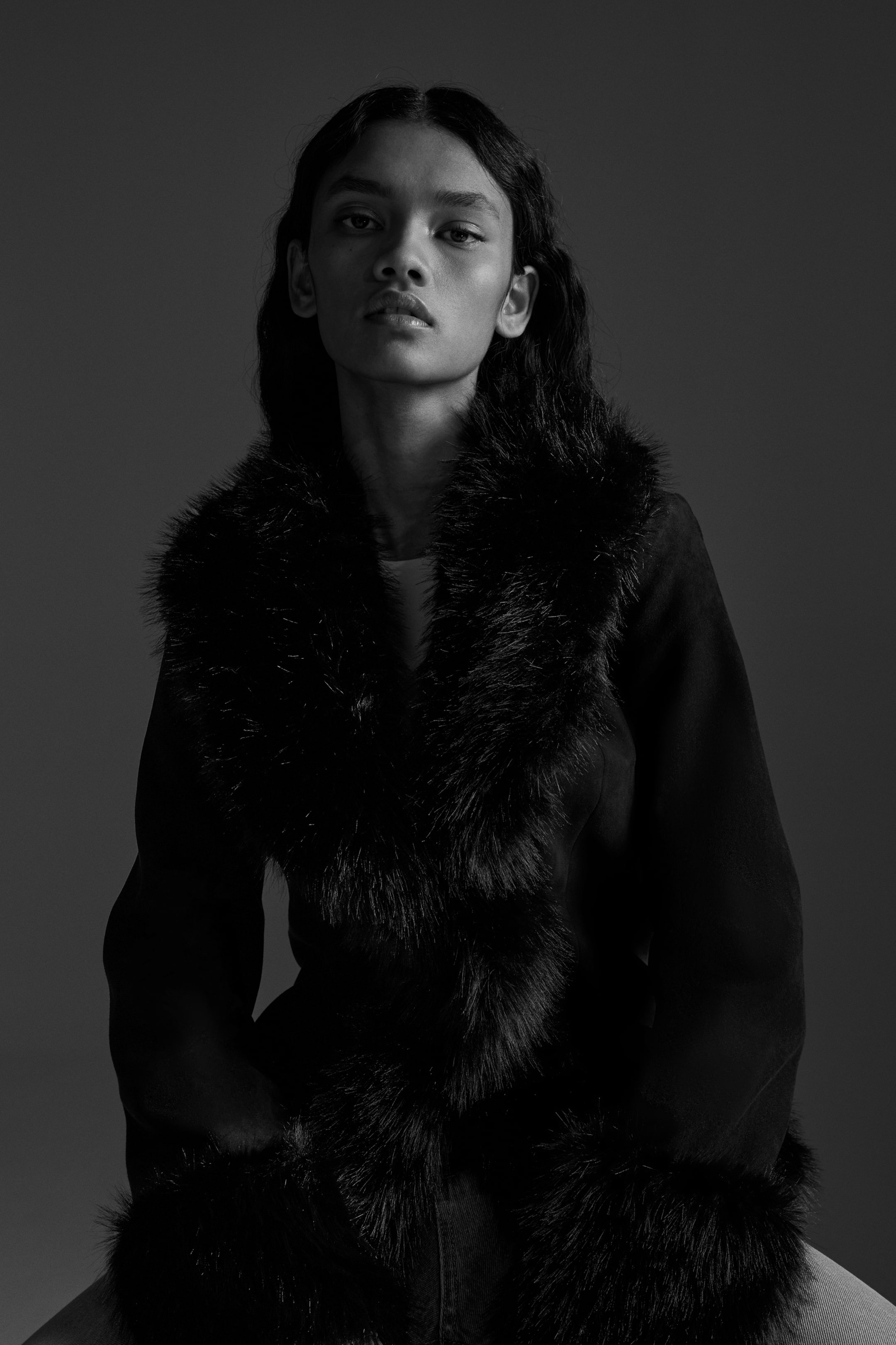 Agrandir l'image: Un manteau sombre est présenté de face, avec une large bordure en fourrure moelleuse. La fourrure longe les revers, descend le long de l'ouverture avant et entoure les poignets, contrastant avec le tissu principal lisse du manteau.