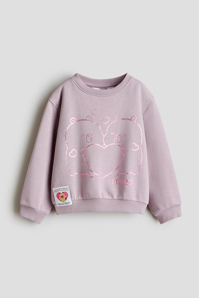A light purple sweatshirt is shown from the front, featuring a round rib-trimmed neckline, dropped shoulders, and long sleeves with ribbed cuffs and hem. The chest displays a shiny pink metallic graphic depicting two cartoon capybaras holding a heart, while a small white character patch is sewn near the bottom hem. - 1