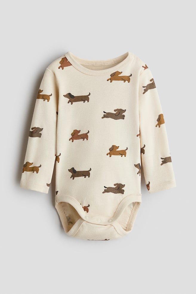 Ein langärmliger Baby-Body aus cremefarbenem Baumwolljersey liegt flach in Vorderansicht und zeigt einen Allover-Print mit Dackeln in Brauntönen. Der Body verfügt über einen Rundhalsausschnitt mit feiner Einfassung, lange Ãrmel und einen Druckknopfverschluss im Schritt. - 1