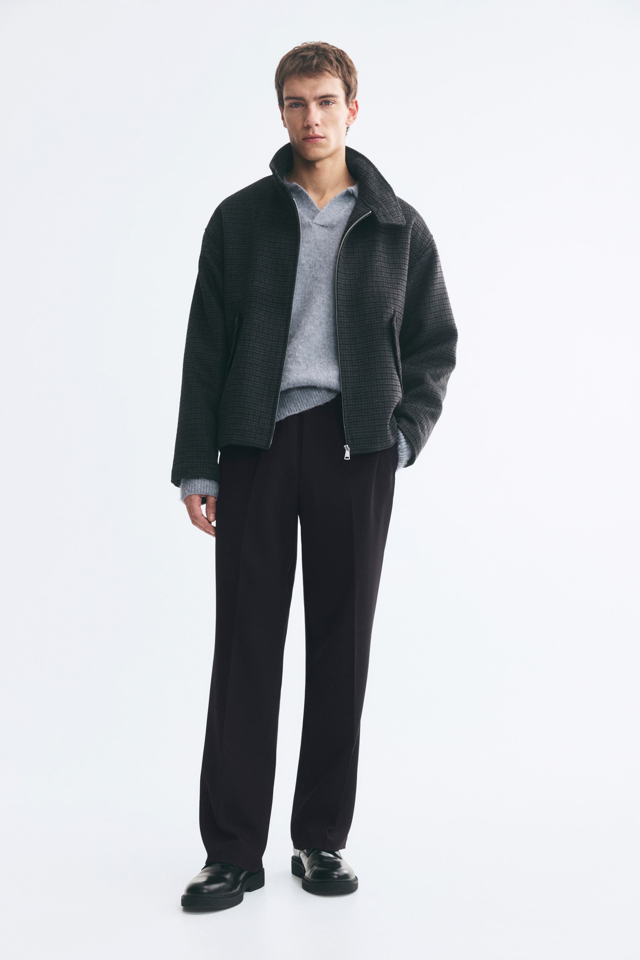 Grotere afbeelding bekijken: Op maat gemaakte joggers van donker geweven stof worden gedragen door het model, met een relaxed silhouet en duidelijke persplooien langs de voorkant van beide pijpen. De broek is gestyled met een lichtgrijze gebreide trui, een donker getextureerd jack met ritssluiting en zwarte leren schoenen.