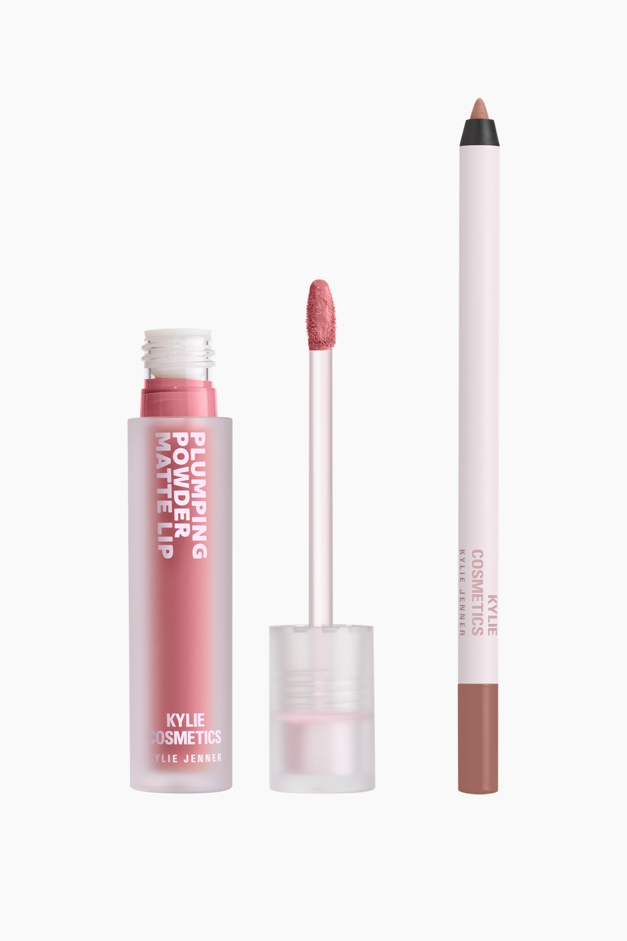 Visa större bild: Kylie Cosmetics Plumping Matte Lip Kit visas med en öppnad, frostig flaska med dammrosa flytande läppstift bredvid en upprätt applicatorstav med en genomskinlig kork. En matchande smal läppenna står vertikalt till höger, med en ljusrosa kropp med varumärkesmärkning och en färgkodad bas.