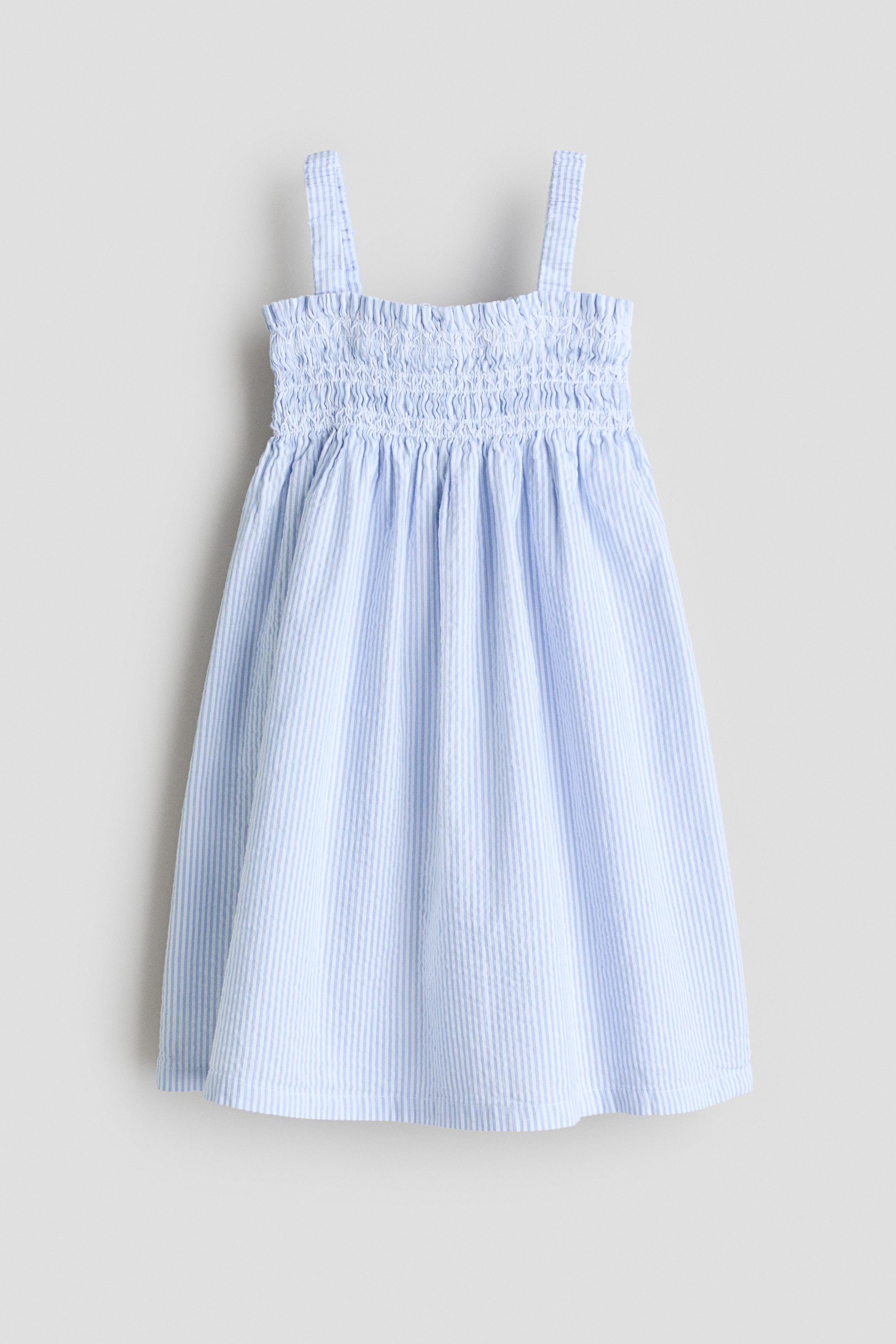 Ver imagem maior: Um vestido está apresentado numa vista frontal, feito de tecido entrançado com riscas verticais em azul claro e branco. Apresenta um decote quadrado com folhos, com smocking em todo o corpete, alças de tecido nos ombros e uma bainha de corte reto.