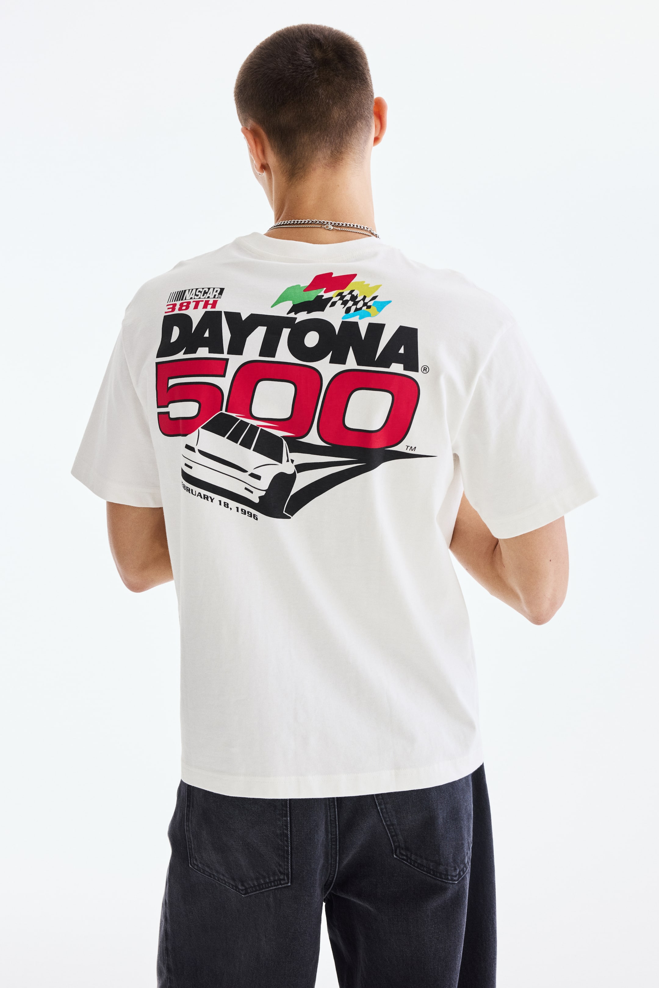 View larger image: The model is wearing a white T-shirt featuring a large, multi-colored graphic print on the back. The print showcases the text "NASCAR 38TH DAYTONA 500," a racing car, and checkered flags. This shirt offers a loose fit with dropped shoulders.