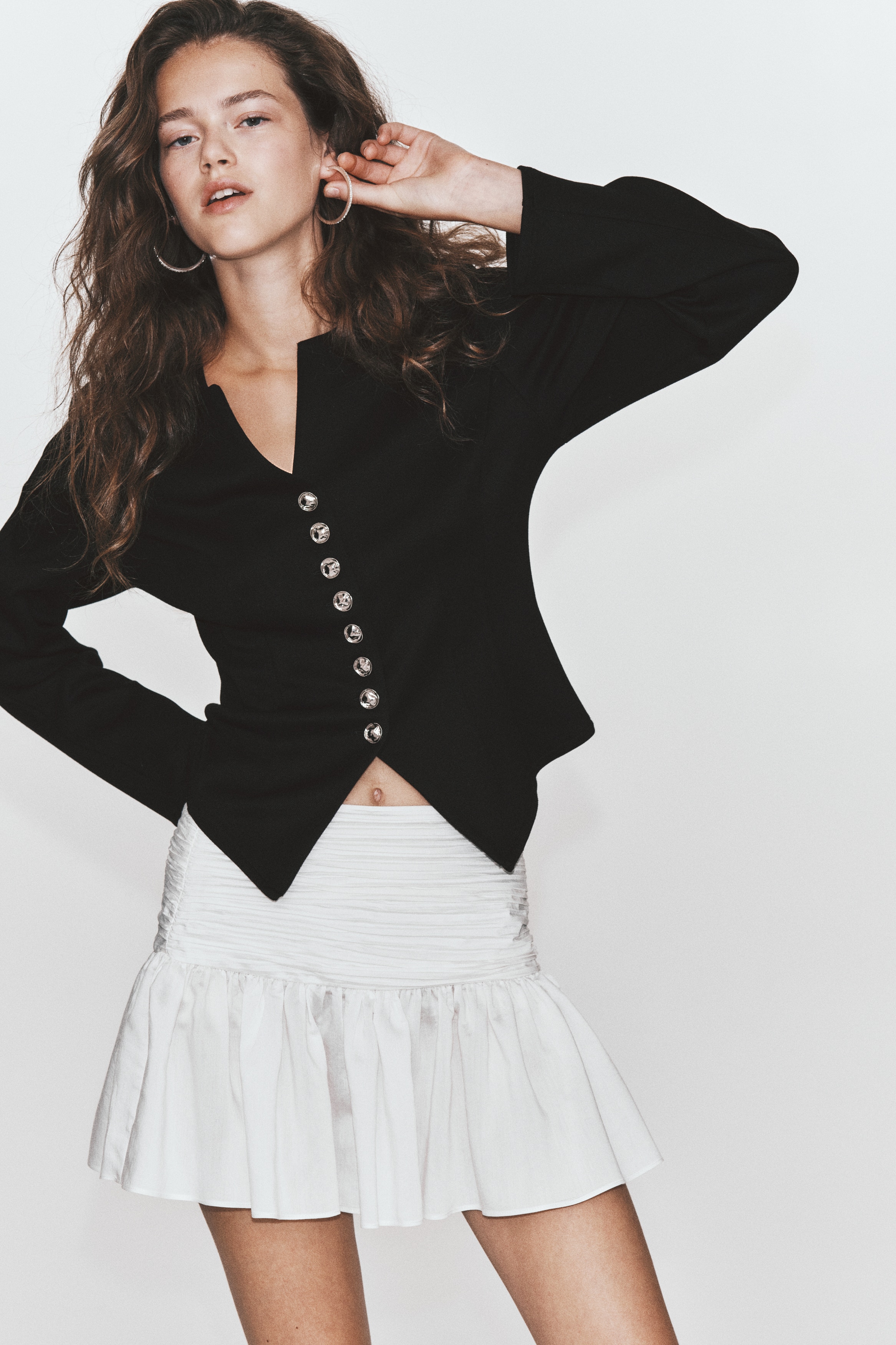 A black, long-sleeved jacket is shown front-facing, featuring a round neckline with a small central V-notch and structured vertical seams along the bodice. It fastens down the centre with a row of eight ornate metallic silver buttons, leading to a pointed, split hem.