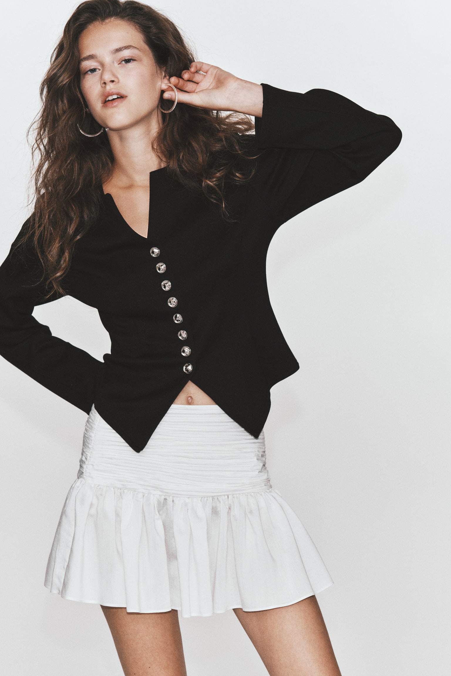 A black, long-sleeved jacket is shown from the front, featuring a round neckline with a small central V-notch and structured vertical seams along the bodice. It fastens at the centre with a row of eight ornate metallic silver buttons and finishes with a pointed, split hem. - 1