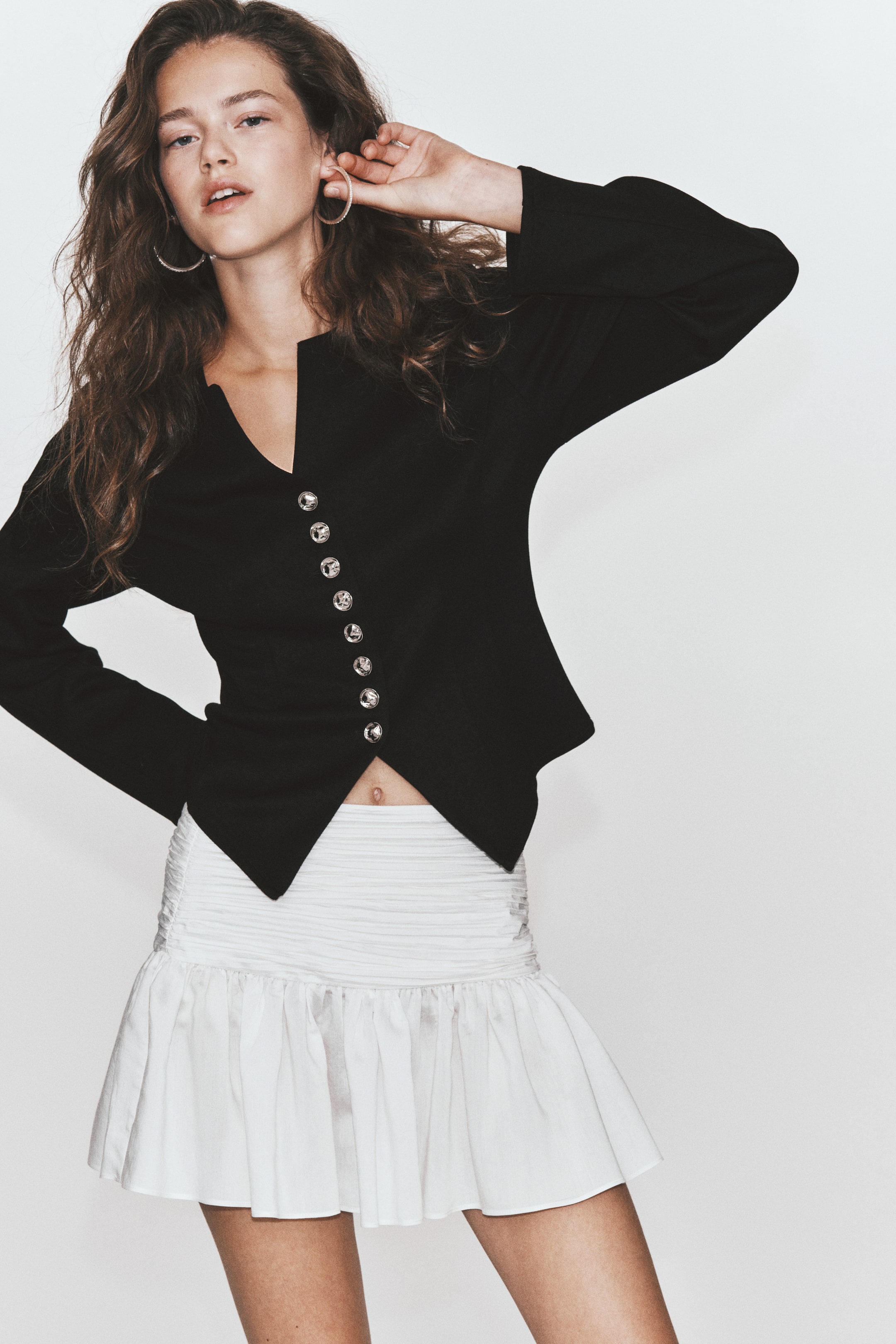 Ampliar la imagen: La modelo lleva un cárdigan negro entallado, combinado con una minifalda blanca con olanes. El cárdigan presenta un escote redondo con estilo abierto en la parte superior, mangas largas y un cierre frontal con botones de tono plateado metálico, dejando los botones inferiores sin abotonar para crear un dobladillo con abertura.