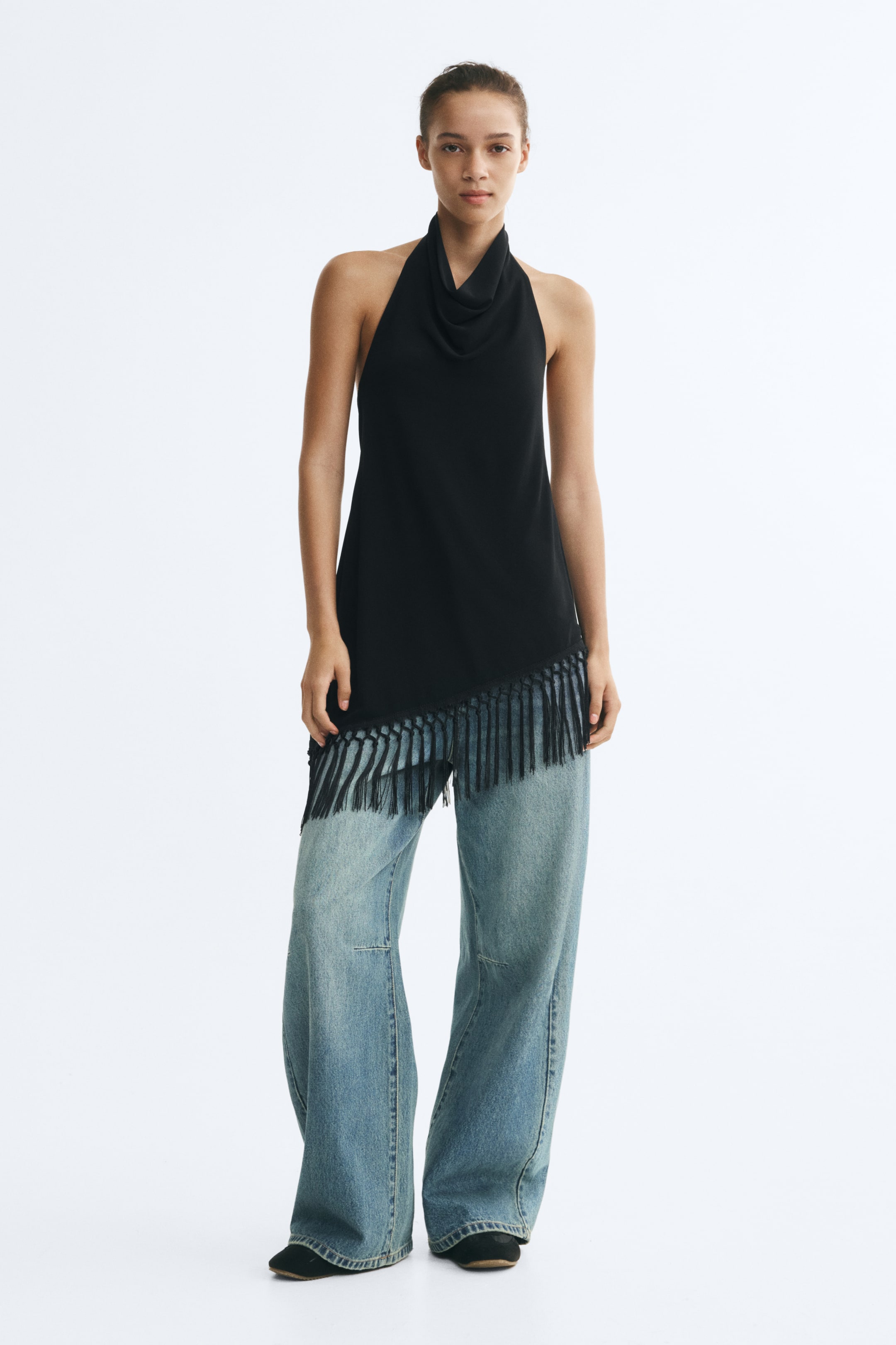 Grotere afbeelding bekijken: Het model draagt een zwarte haltertop met een gedrapeerde halslijn en een asymmetrische zoom met lange franjes en macraméknoopdetails. De top is gestyled met lichtblauwe jeans met wijde pijpen.
