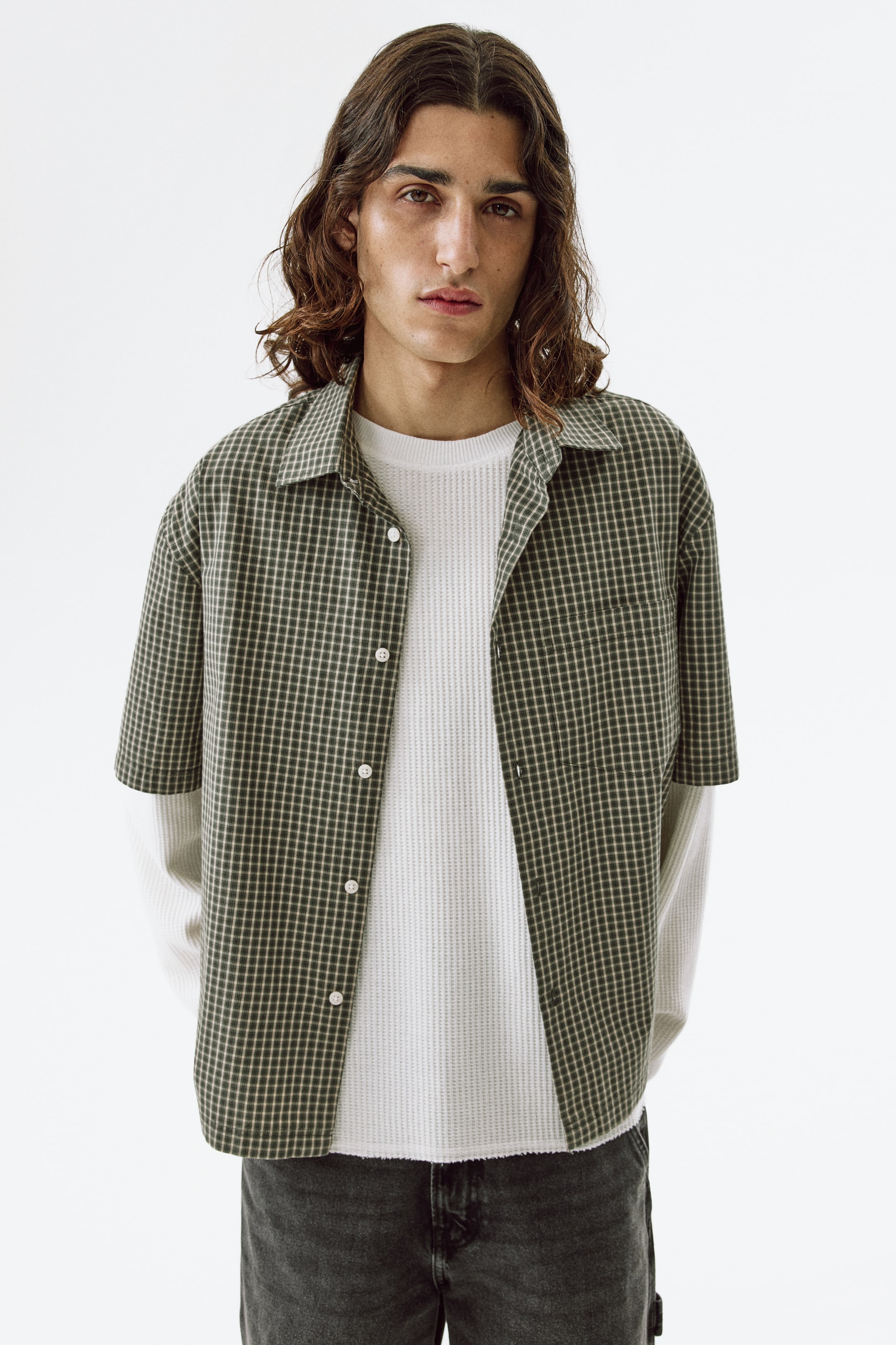 View larger image: Presenting a dark green and beige plaid short-sleeved shirt, featuring a spread collar and a button-down front with white buttons. It includes a left chest patch pocket. The shirt is styled open, layered over a white long-sleeved waffle knit shirt, which showcases a crew neck and a raw hem.