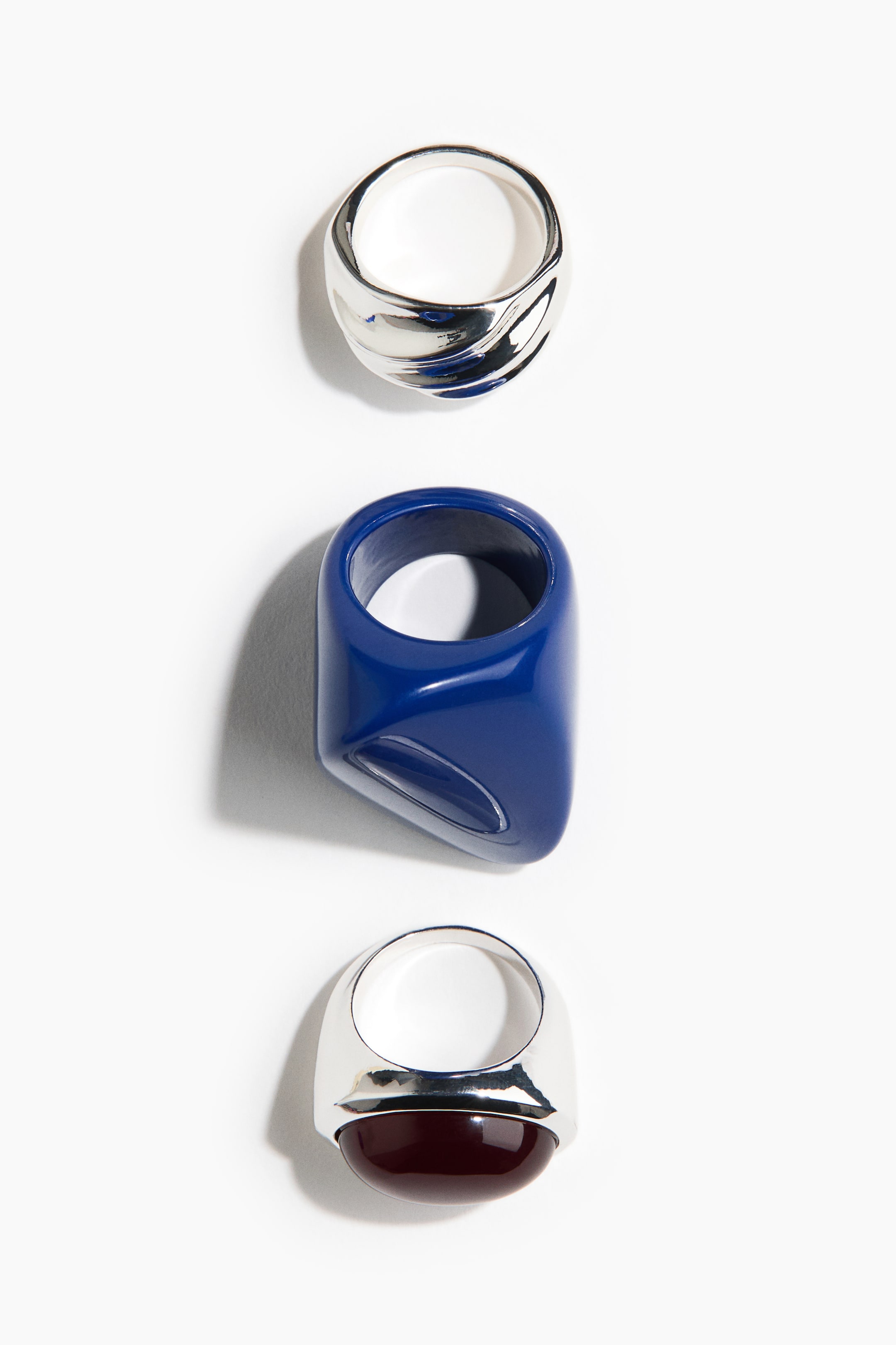Grotere afbeelding bekijken: Een glanzende zilveren ring met een gedraaide, vloeiende band. Een brede, glanzende koningsblauwe ring met een asymmetrische, afgeronde geometrische vorm. Een zilveren ring met een opvallende, gewelfde donkerrode cabochon in de band gezet.