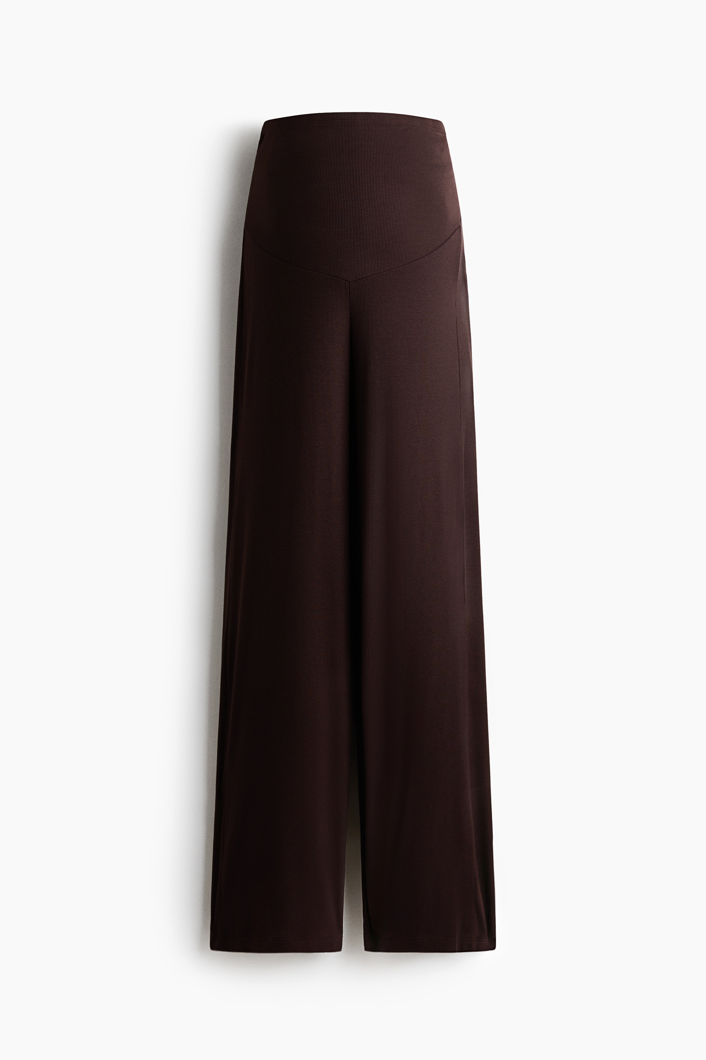 Pantaloni maro închis din jerseu de vâscoză ribat, cu talie înaltă și crac lat, purtați de model dintr-o vedere din spate, din trei sferturi. Pantalonii sunt asortați cu un top alb fără mâneci, o jachetă neagră purtată pe umeri și pantofi sport maro.