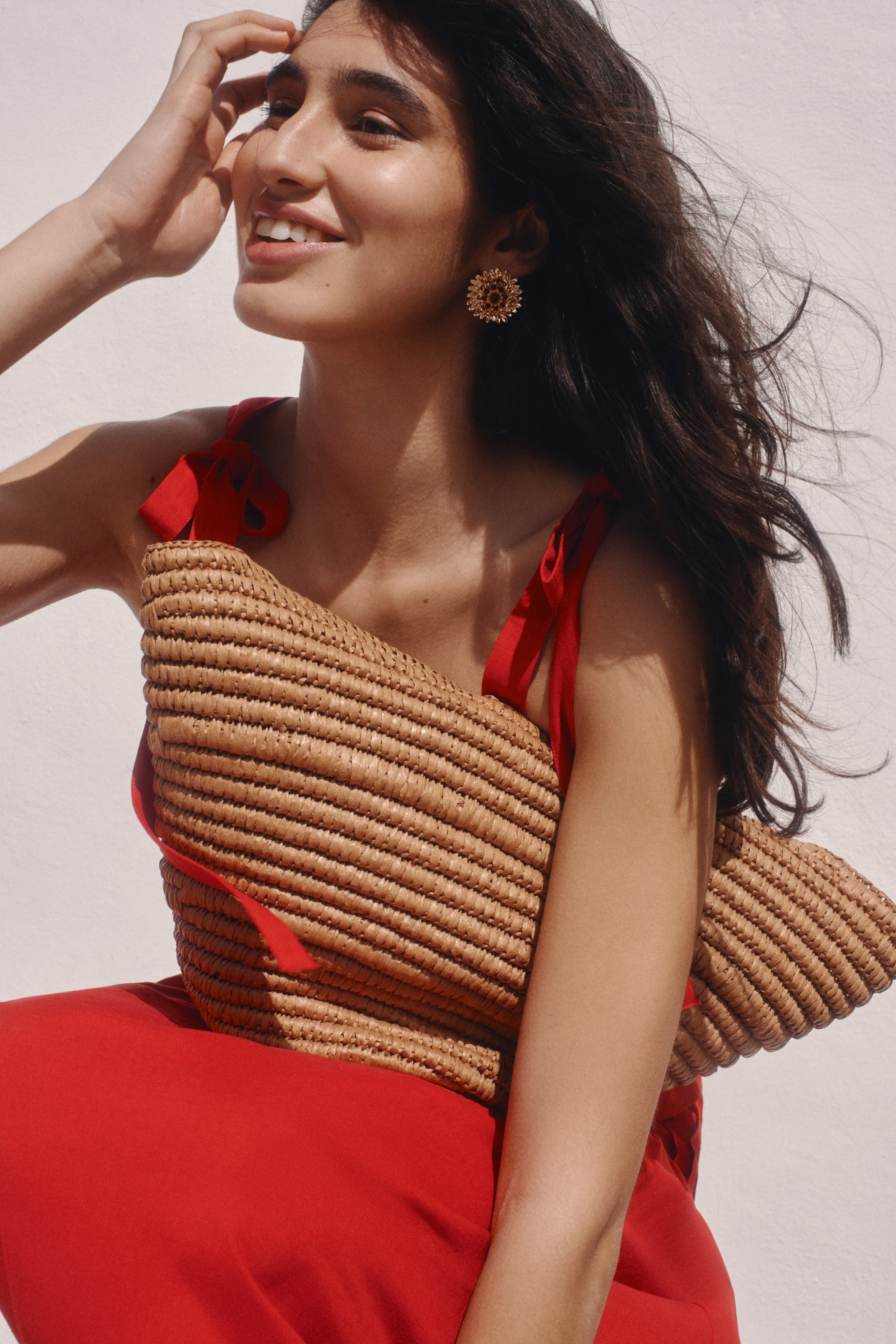 View larger image: A natural straw woven bag is being held under the right arm, angled forward and slightly downward. It features a rectangular shape and a tightly woven horizontal pattern, set against a red garment.