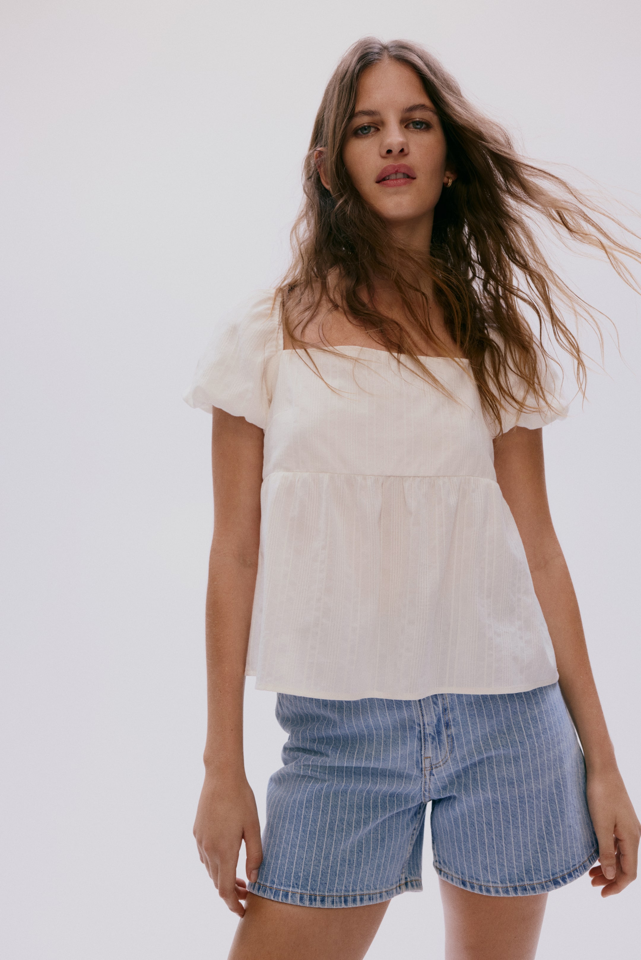 Ampliar la imagen: Un top blanco roto con un escote cuadrado, mangas cortas abullonadas y una textura sutil de rayas verticales, con un dobladillo peplum fruncido que se forma debajo del busto.